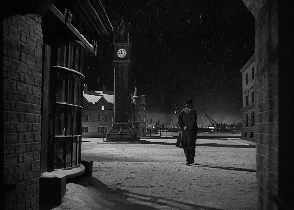 A man in a long dark coat walks through a quiet city square next to a clocktower at night.