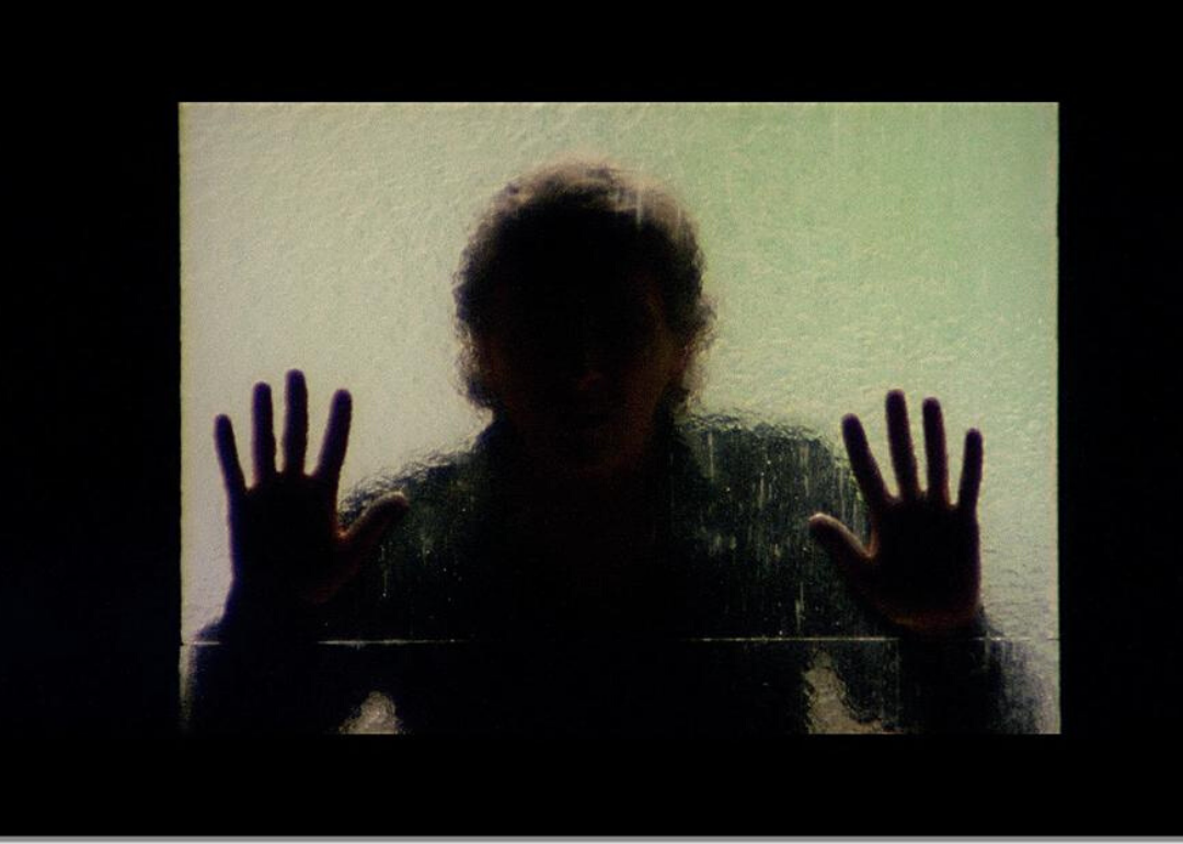 A man stands behind frosted glass holding both hands up.
