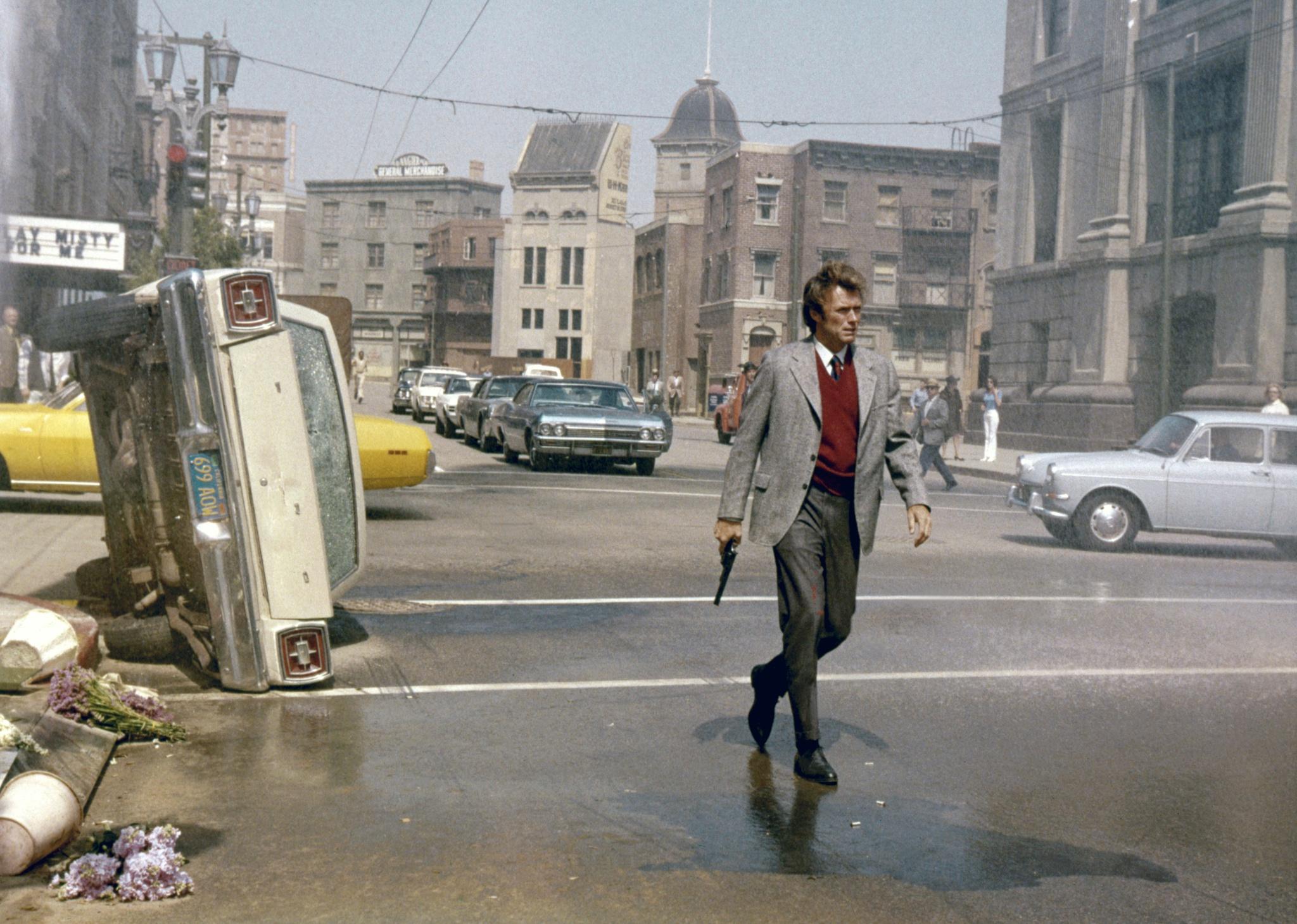 Clint Eastwood, wearing a sweater vest and blazer, walks down a wrecked city street with a gun by his side.