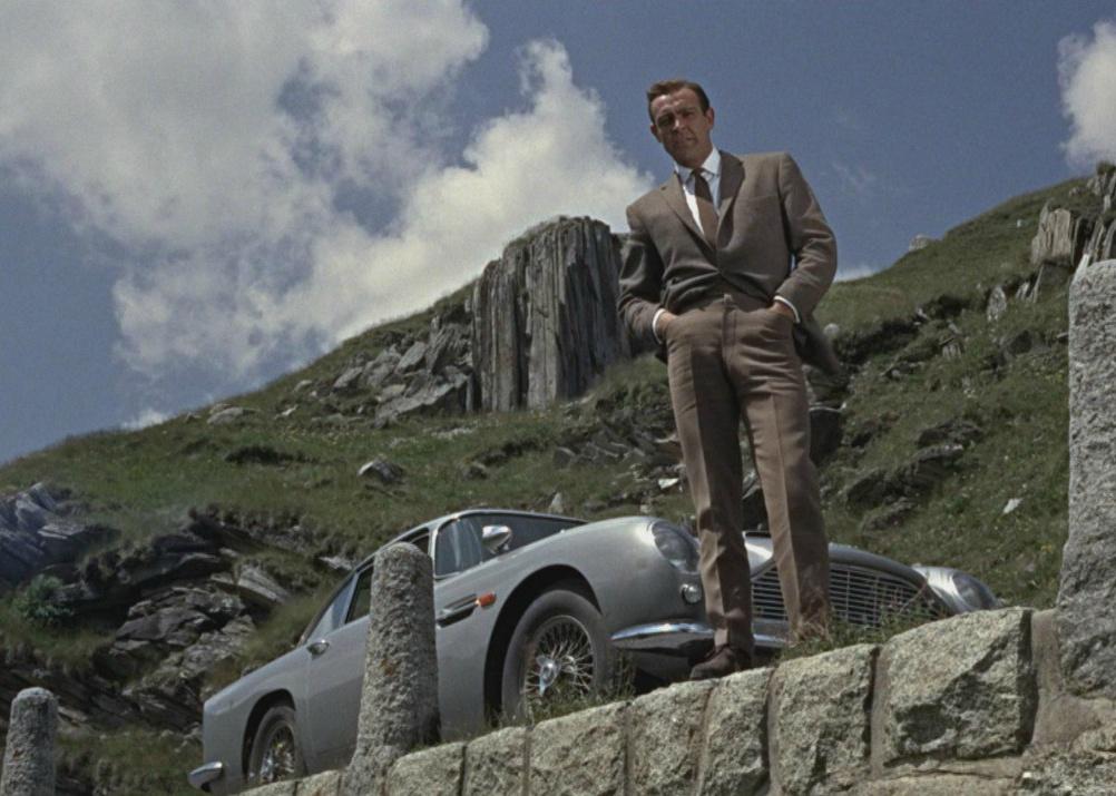 Sean Connery standing on a mountain road next to a classic sports car looking down at something.
