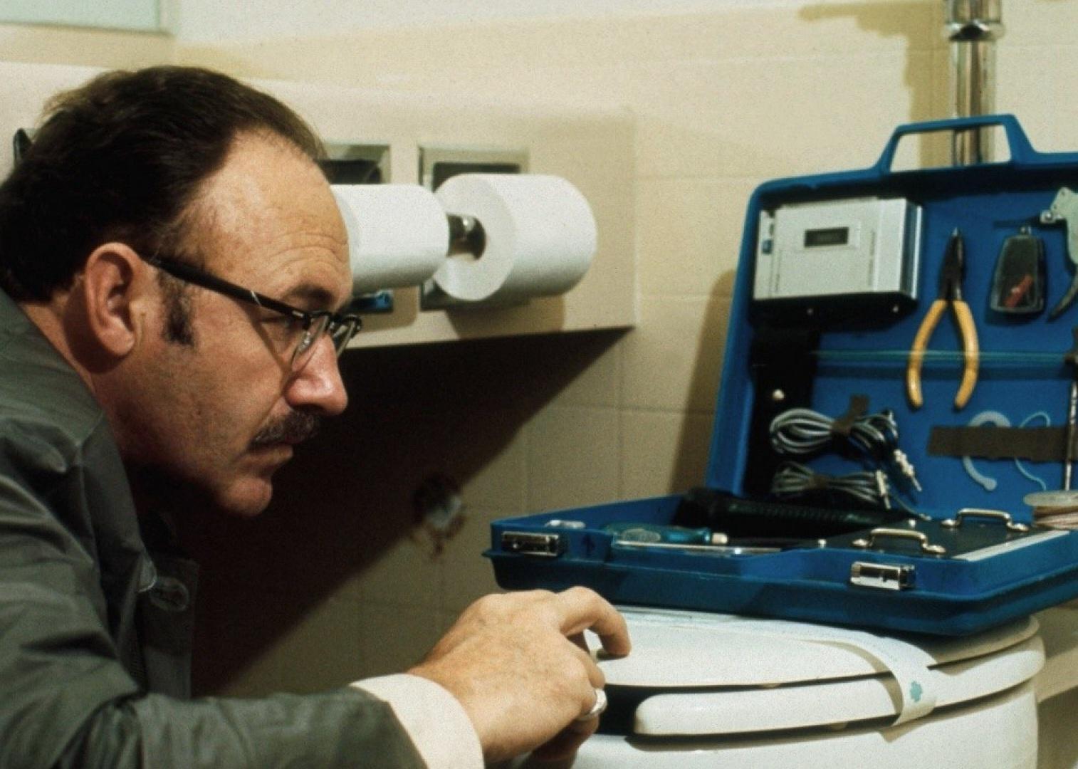 Gene Hackman wearing a suit and glasses, doing something to a toilet with a toolbox.