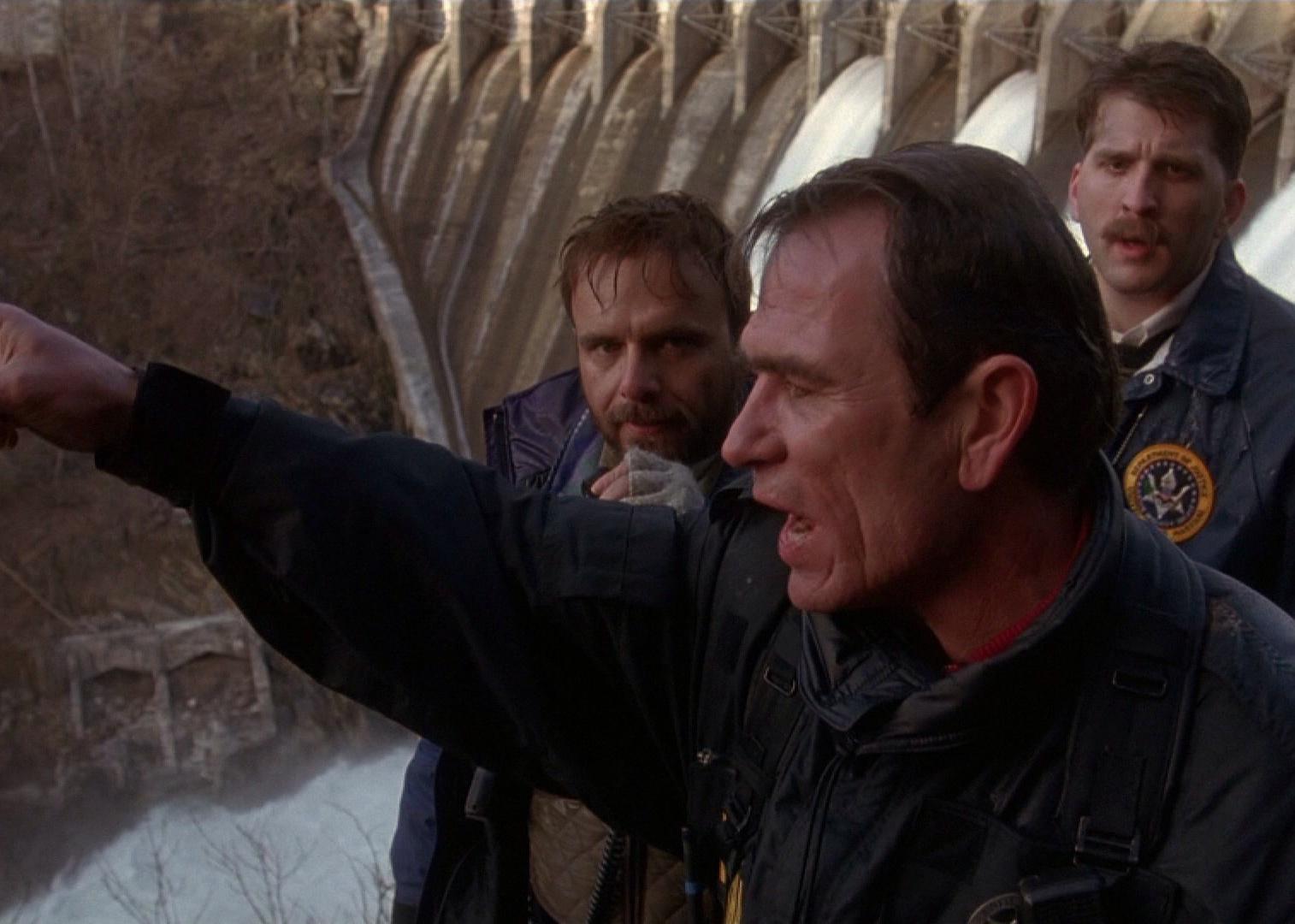 Tommy Lee Jones points at something at the bottom of a dam with Joe Pantoliano and Daniel Roebuck as police officers in the background.