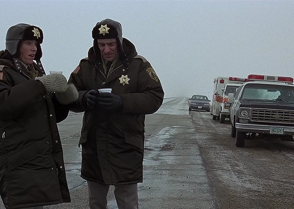 Frances McDormand and another police officer stand outside in the snow sipping coffee and talking next to a scene.