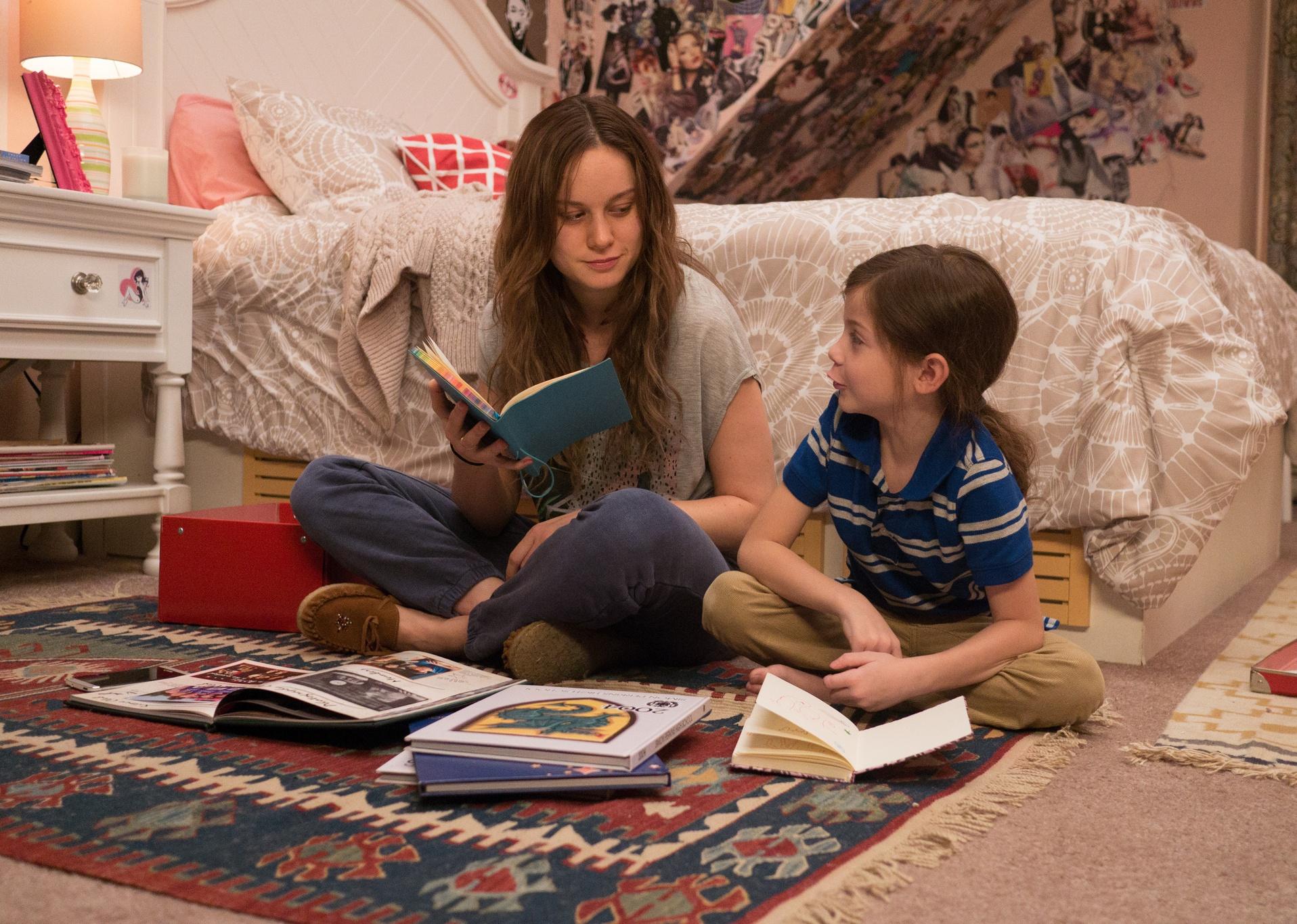 Brie Larson and a young boy sit on the floor reading together in a small bedroom.