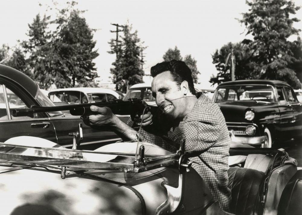A man pointing a long gun from a classic convertible car in black and white.