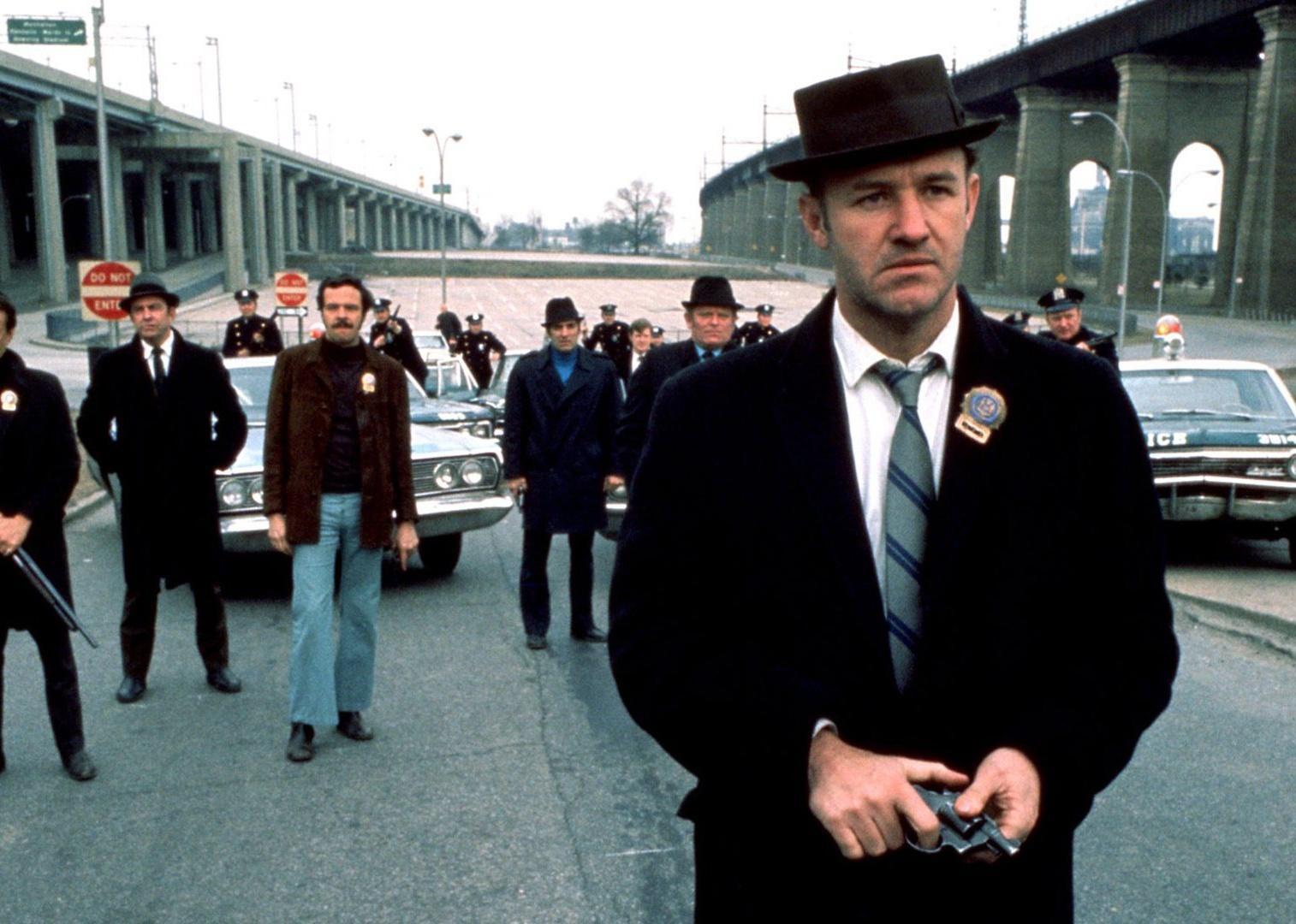 Gene Hackman leads a team of policemen standing in front of police cars.