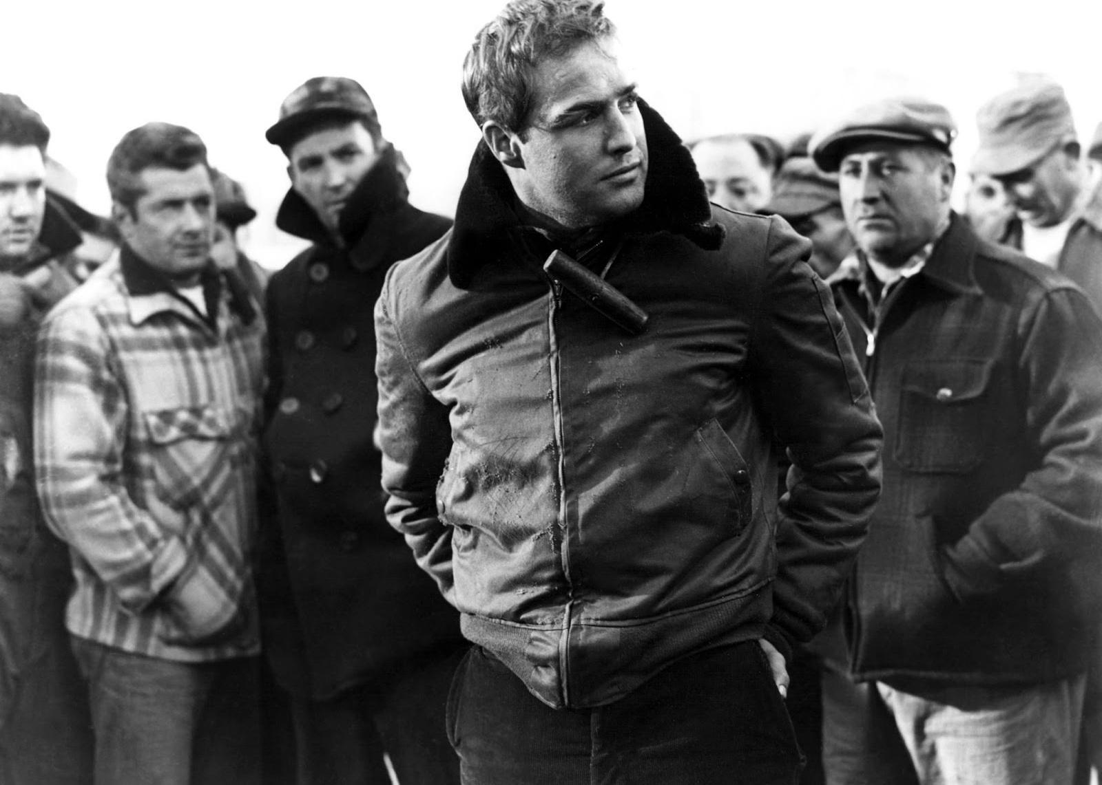 Marlon Brando, wearing a leather jacket, stands in front of a crowd of men.