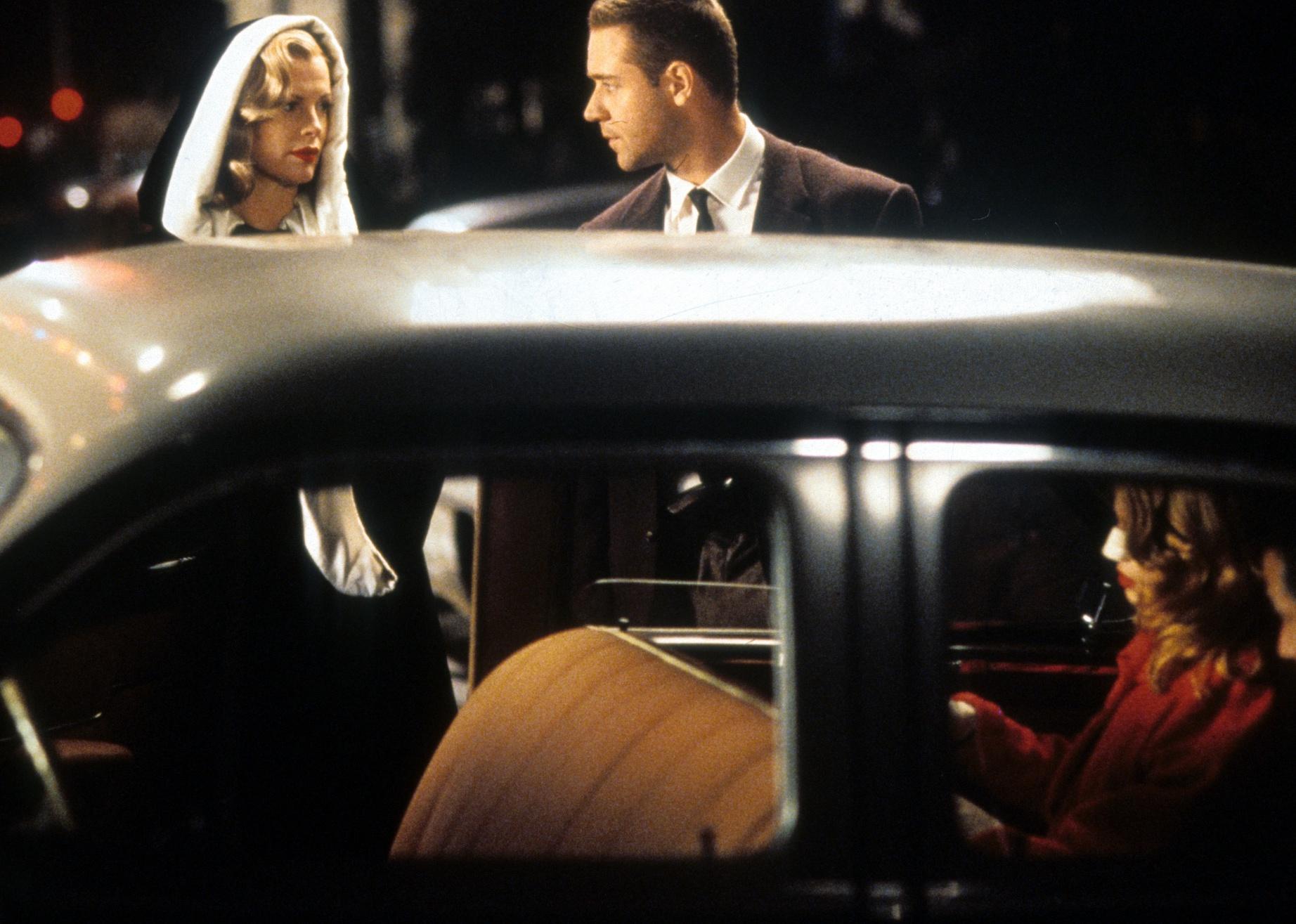 Kim Basinger and Russell Crowe stand dressed up and talking outside a classic car.