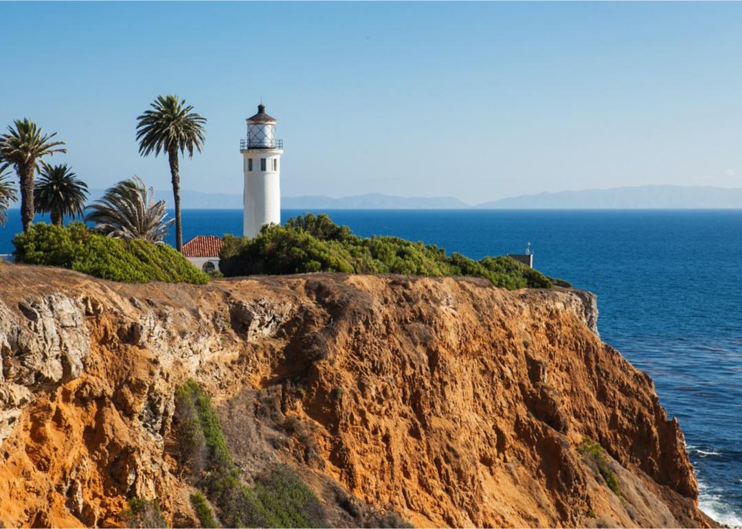 A lighthouse and palm trees on a rocky cliff over the water.