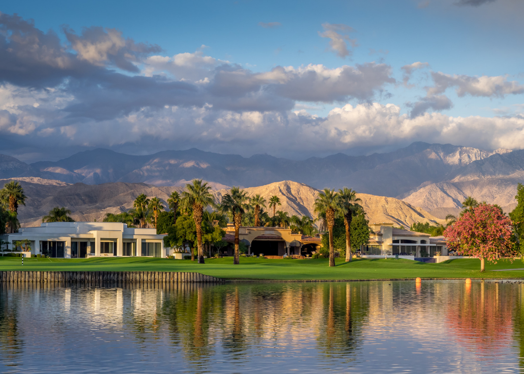 A rolling green lawn on the water in front of a large home with palm trees and mountains in the background.