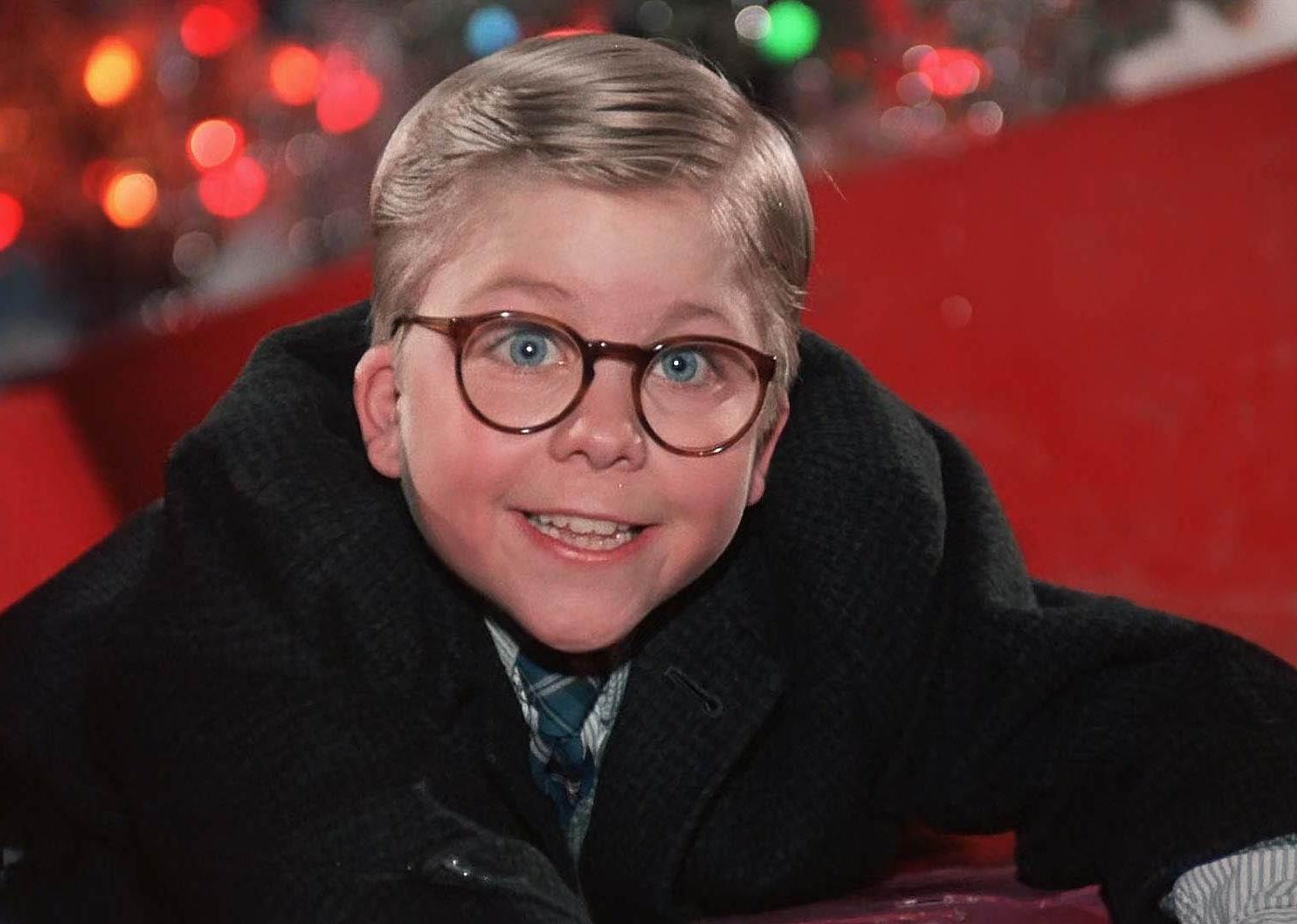 Peter Billingsley as a child smiling and wearing glasses in front of Christmas lights.