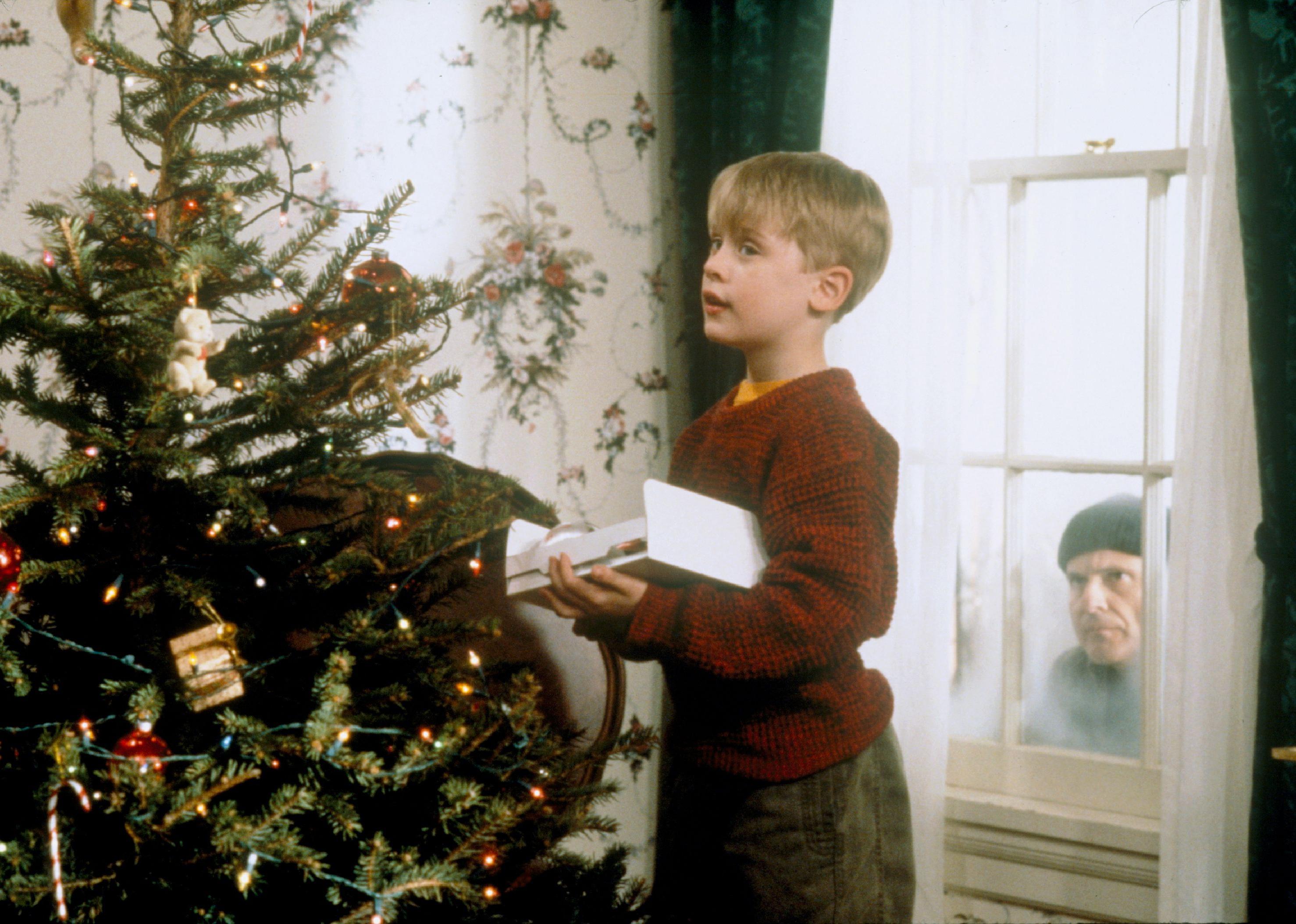Macaulay Culkin decorating a Christmas tree while Joe Pesci peers into the window from outside. 