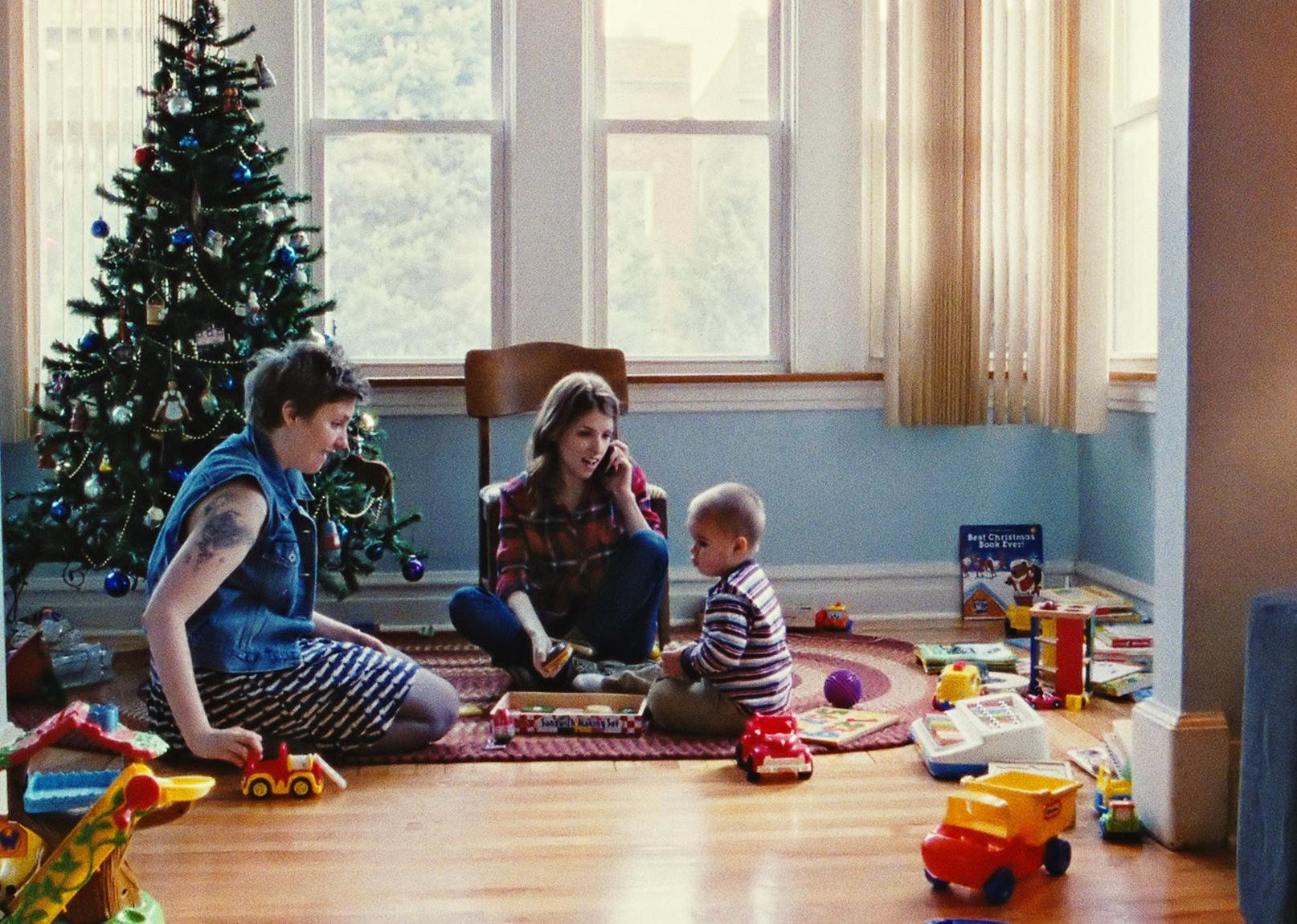 Anna Kendrick and Lena Dunham sitting on the floor in front of a Christmas tree playing with a little boy.
