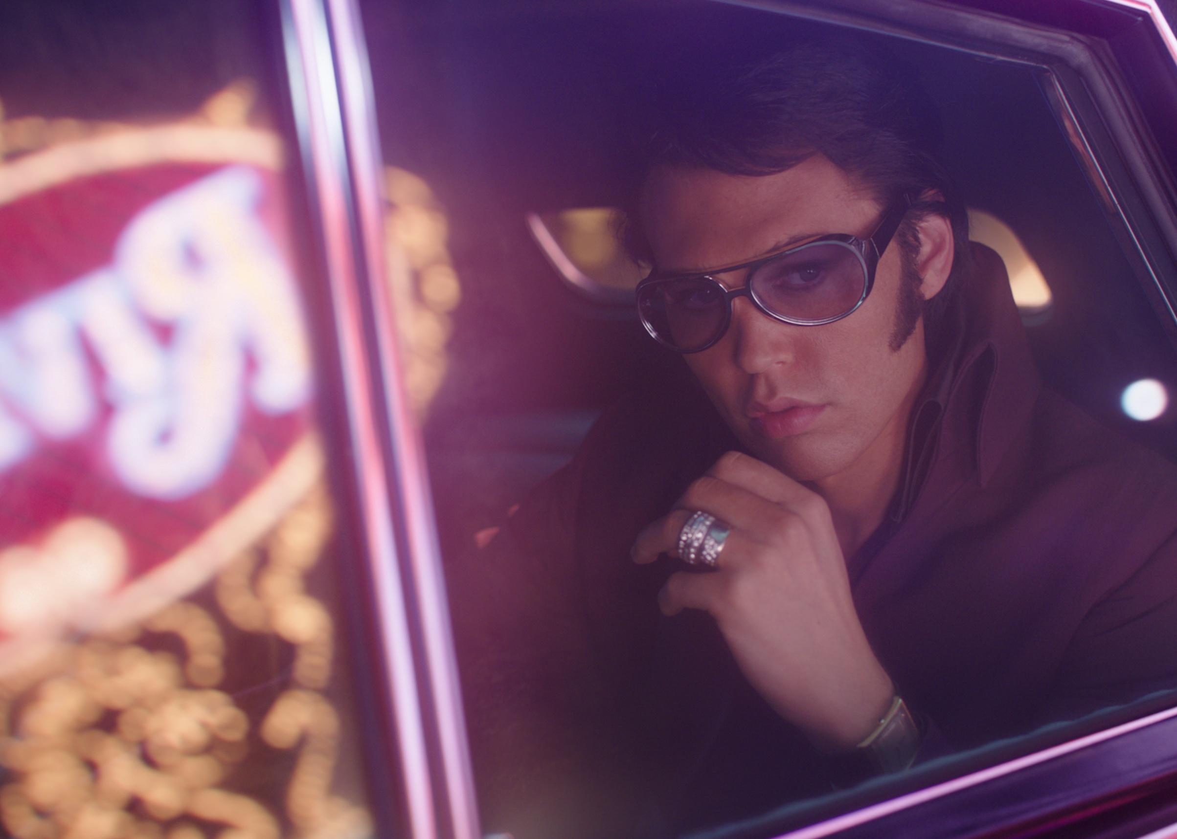 An actor playing Elvis in the backseat of a car with neon signs in the reflection of the car window.