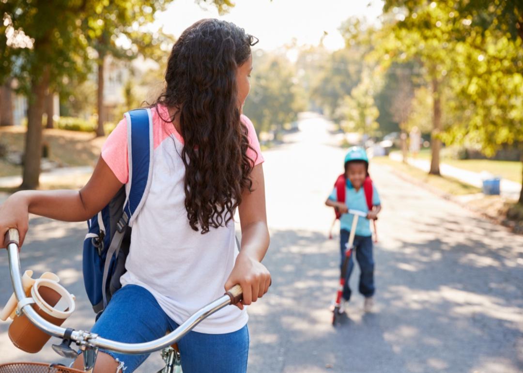 A girl on a bicycle looks back and waits for a little boy on a scooter.