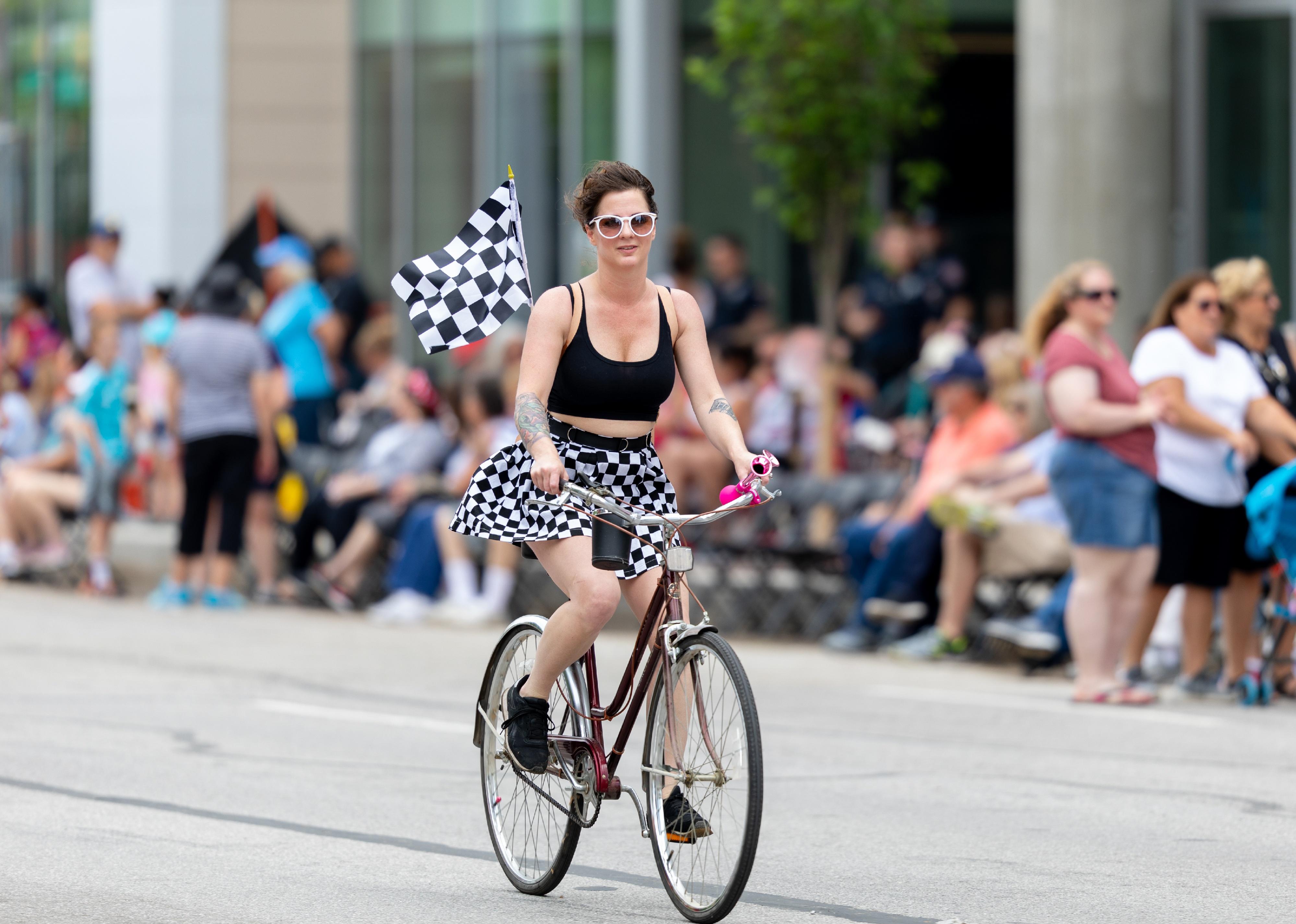 A woman in black and white with a checkered skirt and flag on her bicycle riding down a crowded street.