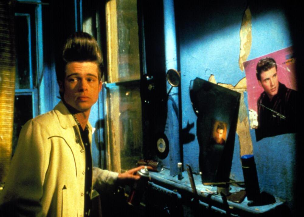 Brad Pitt with a can of spraypaint standing by a blue wall with a large Elvis pomp hairdo.
