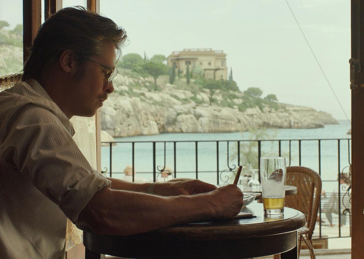 Brad Pitt sitting at a desk, with a view of the sea, writing.