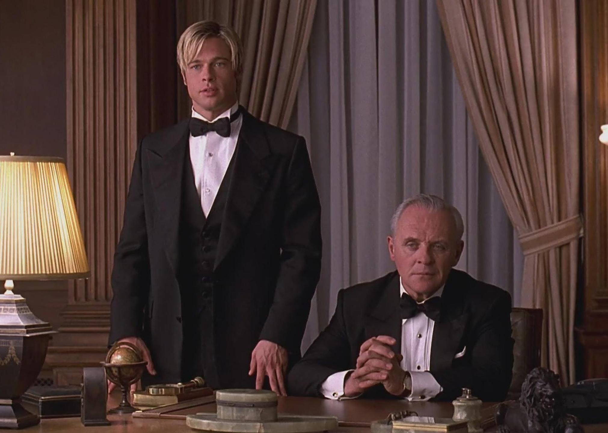 Brad Pitt standing next to Anthony Hopkins, who is seated at a desk, both dressed in suits and bowties.