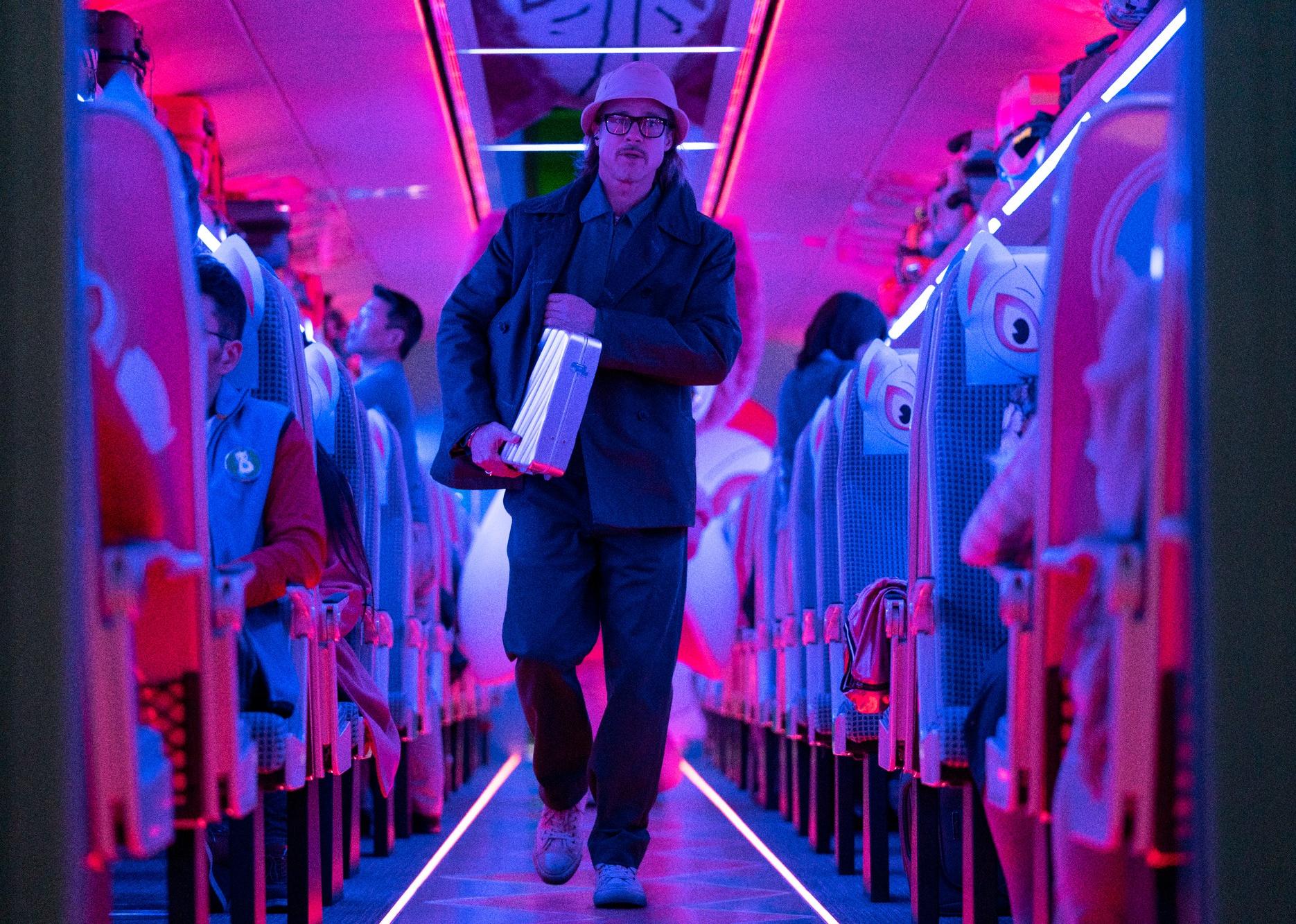 Brad Pitt walks through a crowded train in a bucket hat carrying a metal briefcase under blue and red lighting.