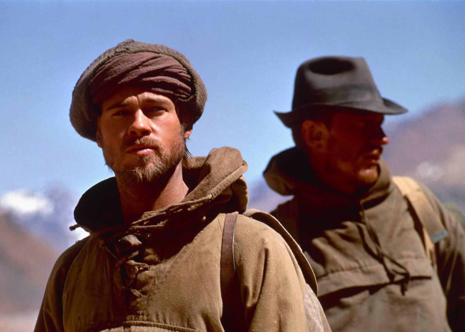 Brad Pitt and David Thewlis hiking in dark green coats and head coverings in the mountains with beards.