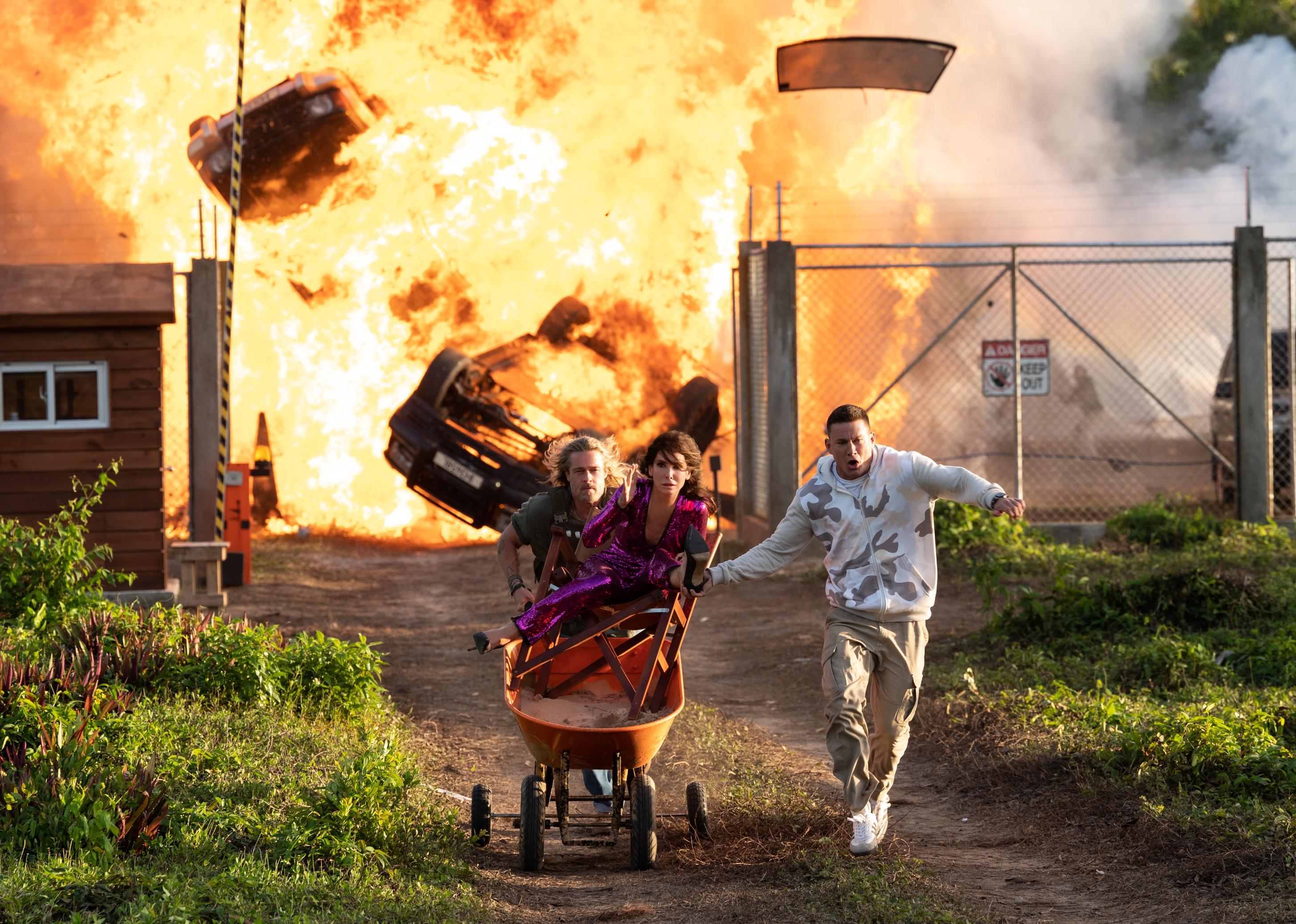 Brad Pitt and Channing Tatum pushing Sandra Bullock in a wheelbarrow from a large explosion in the background.