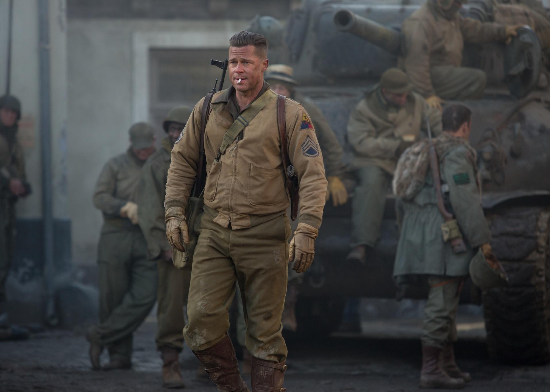 Brad Pitt in 1940s army gear with a gun on his shoulder smoking a cigarette in front of a tank and troops.