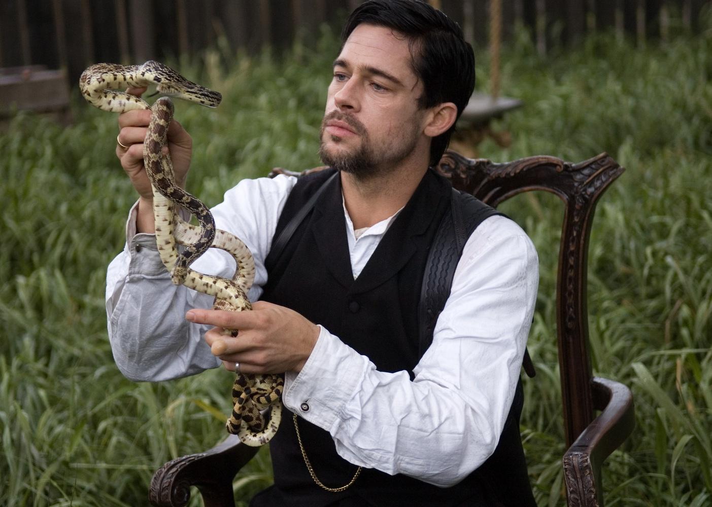 Brad Pitt in a Western shirt and vest sitting in a wooden chair outside holding a snake.