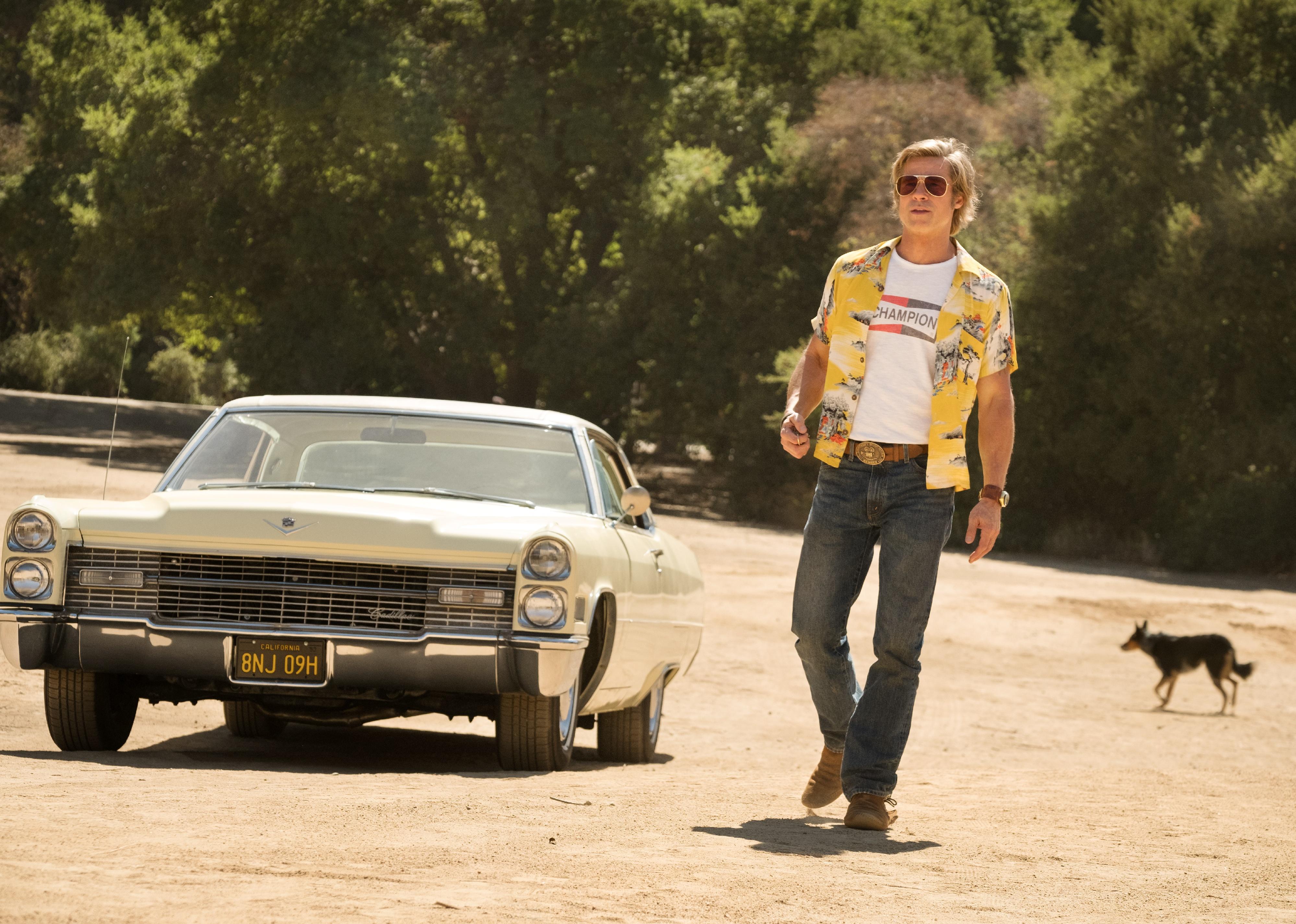 Brad Pitt, wearing jeans, a Western belt buckle, t-shirt and Hawaiian shirt, walking from a classic car.