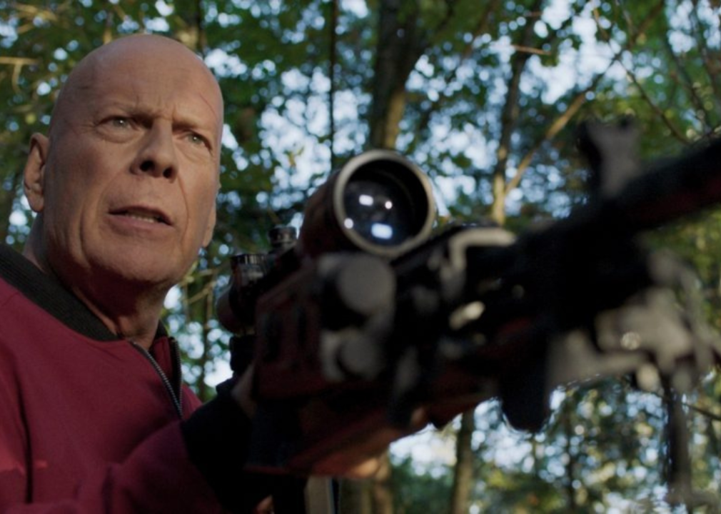 Bruce Willis looking ahead while holding a large gun with a scope.