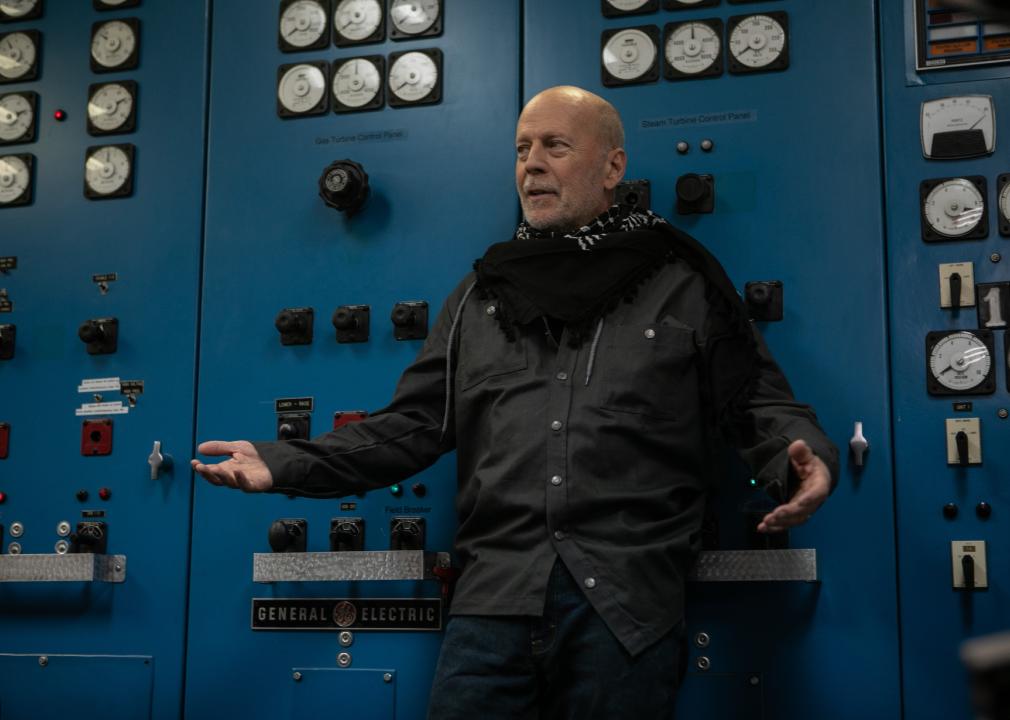 Bruce Willis stands in front of a blue wall of electrical switches.