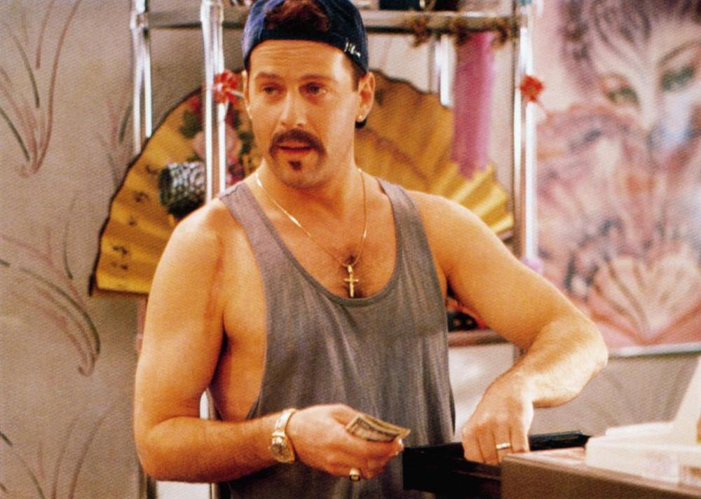 Bruce Willis with cash in hand behind a register dressed in a muscle tank, gold cross necklace and backwards hat.