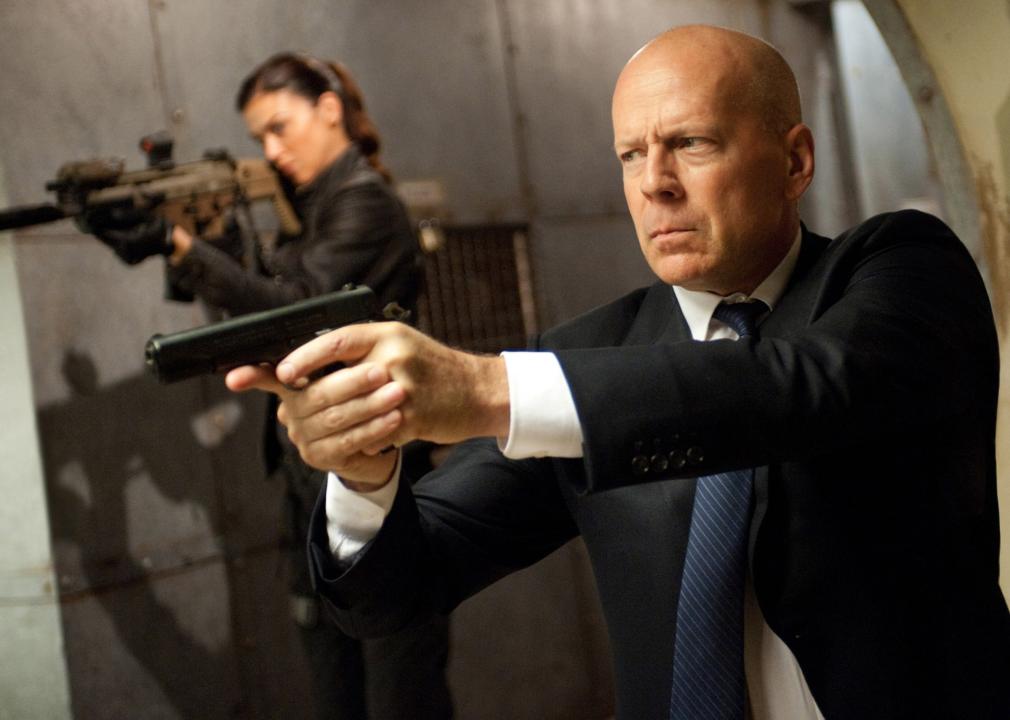 Bruce Willis points a handgun in the foreground while a woman in the background points a larger gun in the same direction.