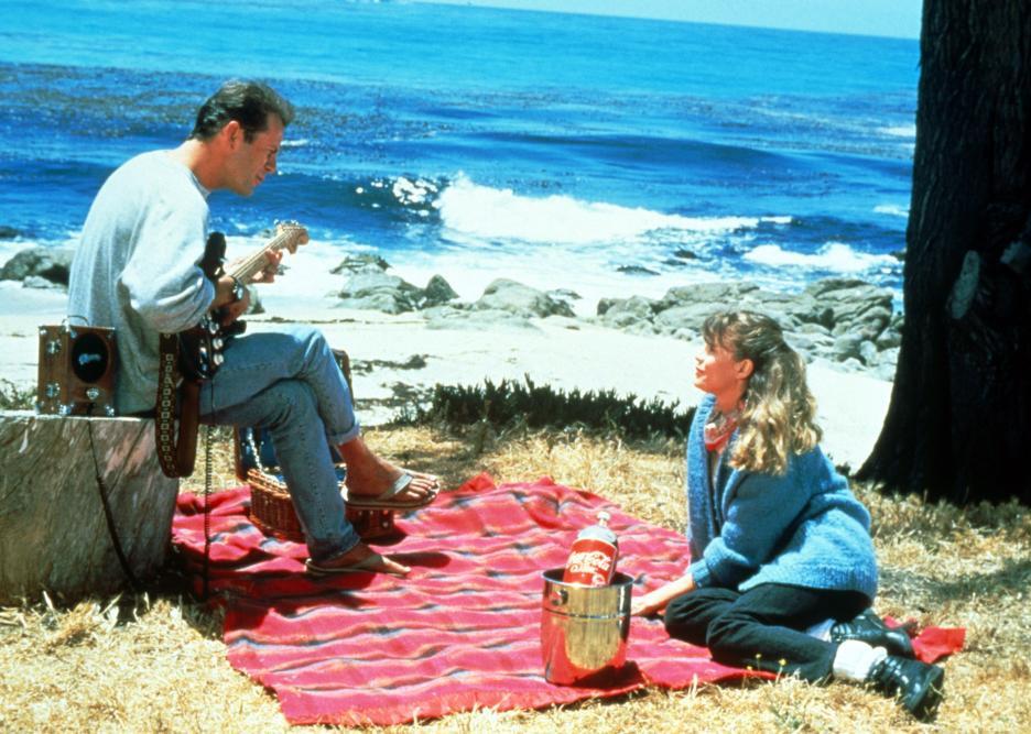 A man plays guitar for a woman sitting on a picnic blanket at the beach.