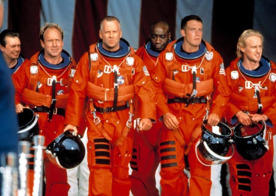 A row of astronauts in orange suits walk together with helmets in hand.