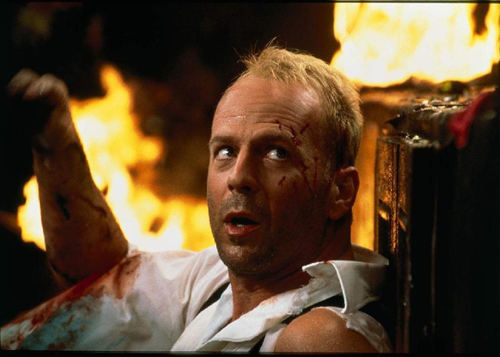 Bruce Willis sits wearing a tattered shirt with fire in the background.