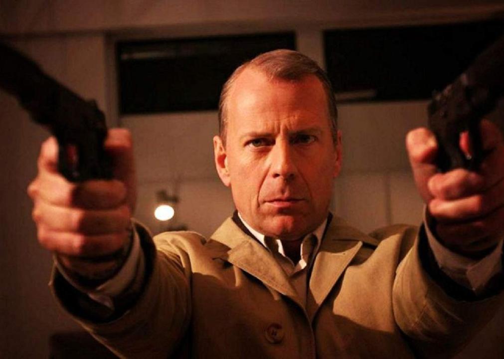 Bruce Willis in a tan trenchcoat shooting a gun in each hand.