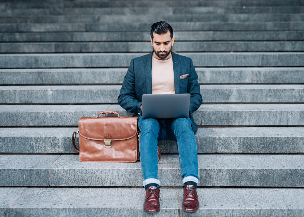 A man with a leather briefcase sitting on steps while working on his laptop.