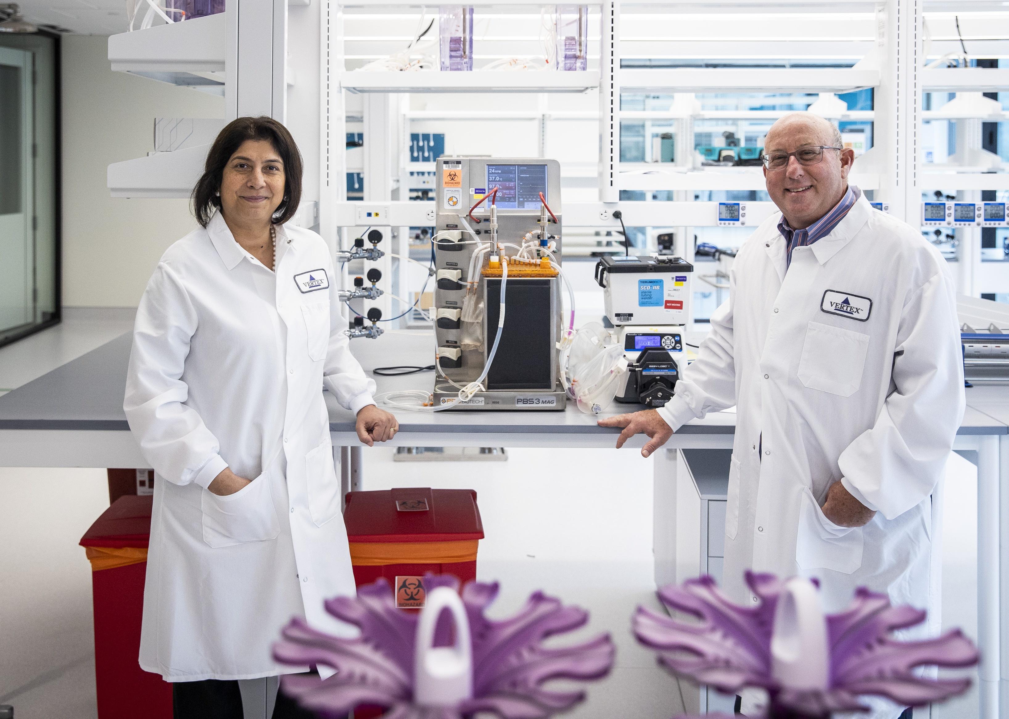 Reshma Kewalramani, left and Jeff Leiden, both wearing white lab coats, pose for a portrait with one of their bioreactors.