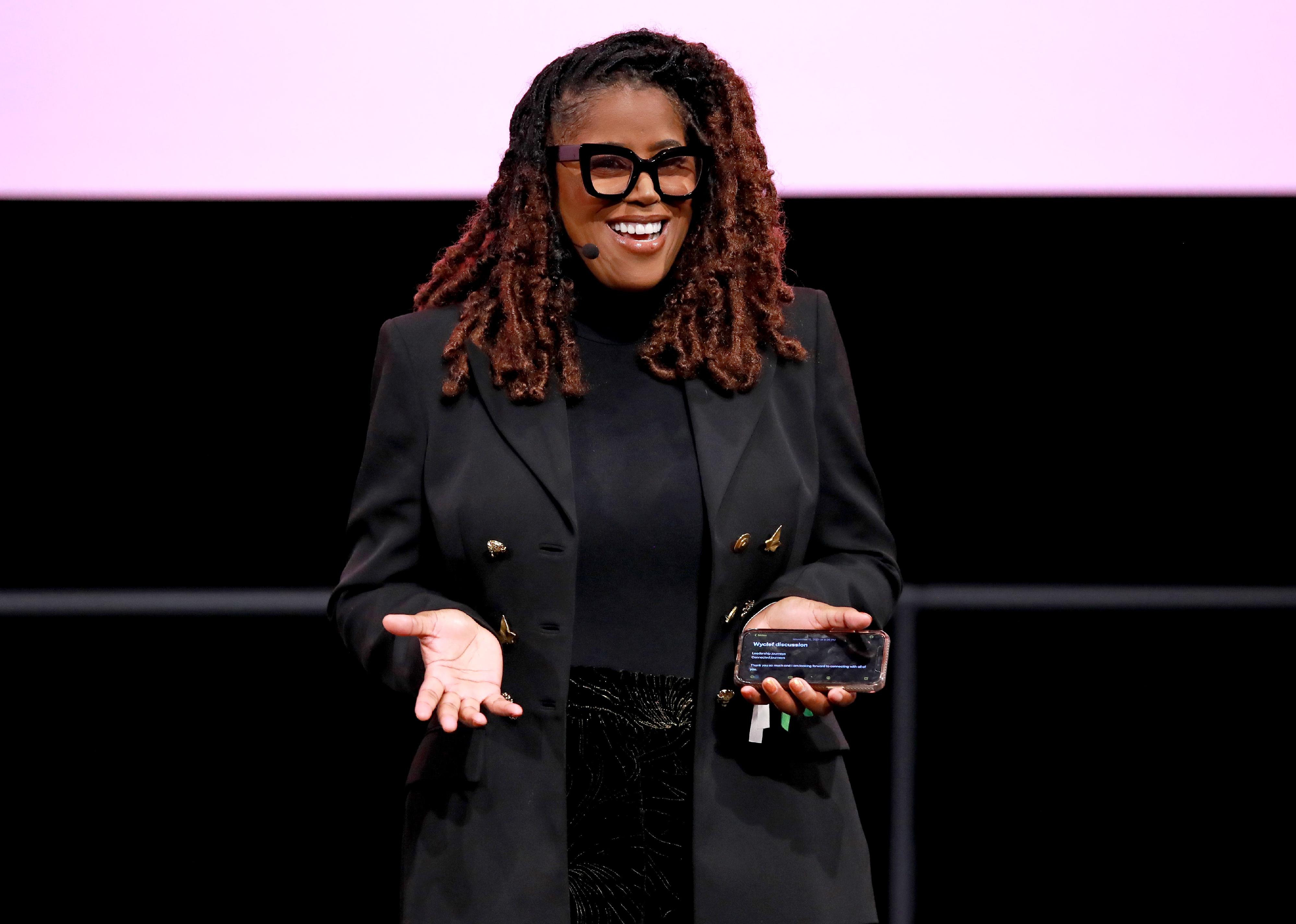 Thasunda Duckett, wearing all black, speaking onstage against a black and pink backdrop.