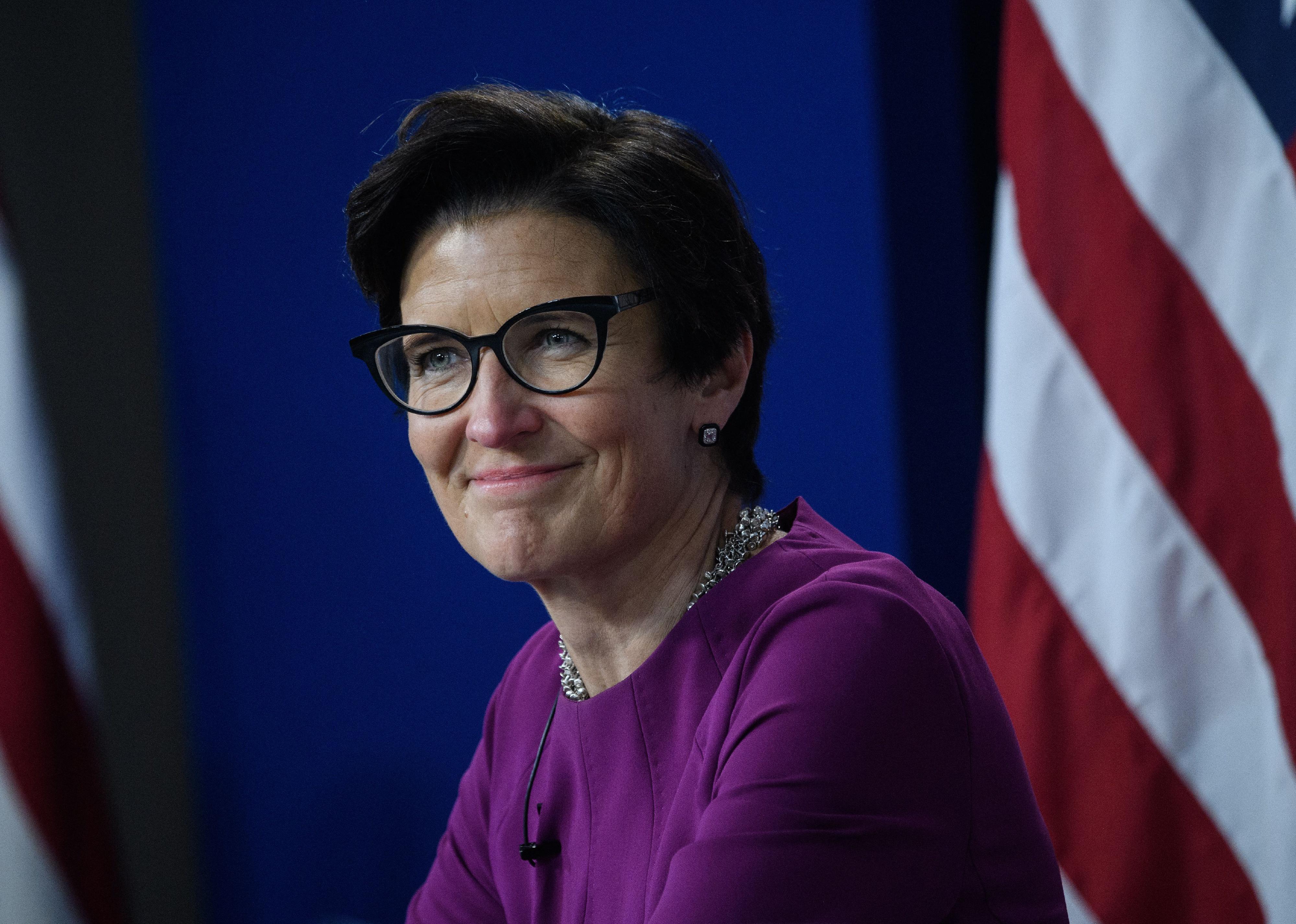 Jane Fraser, in a purple dress, sits smiling in front of a blue background and an American flag.