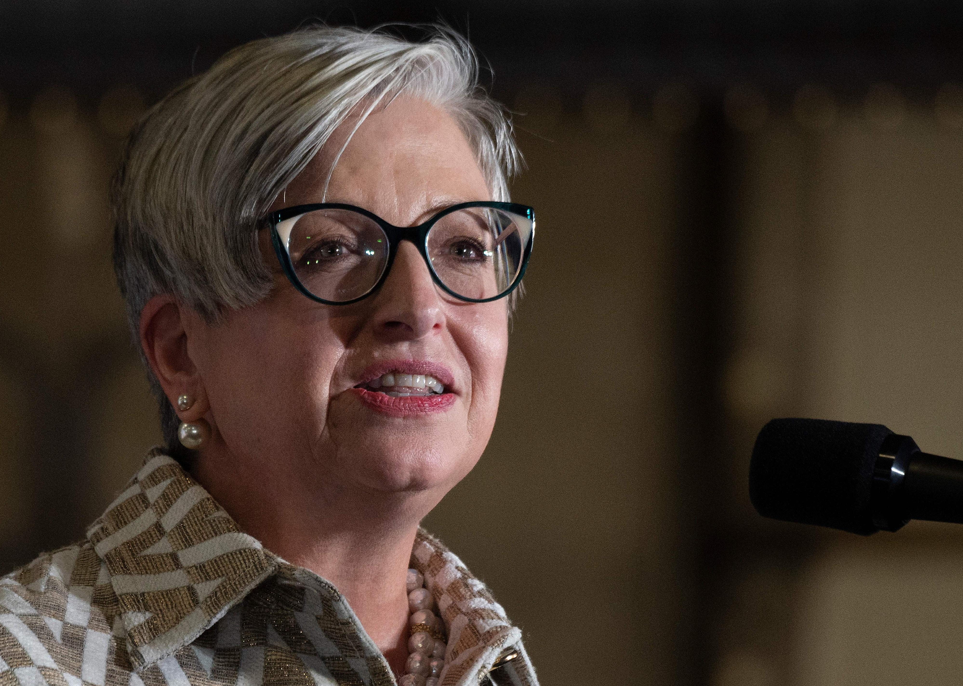Carol Tomé, wearing pearl earrings, a gold and white patterned top and dark framed round glasses, speaking at an event.