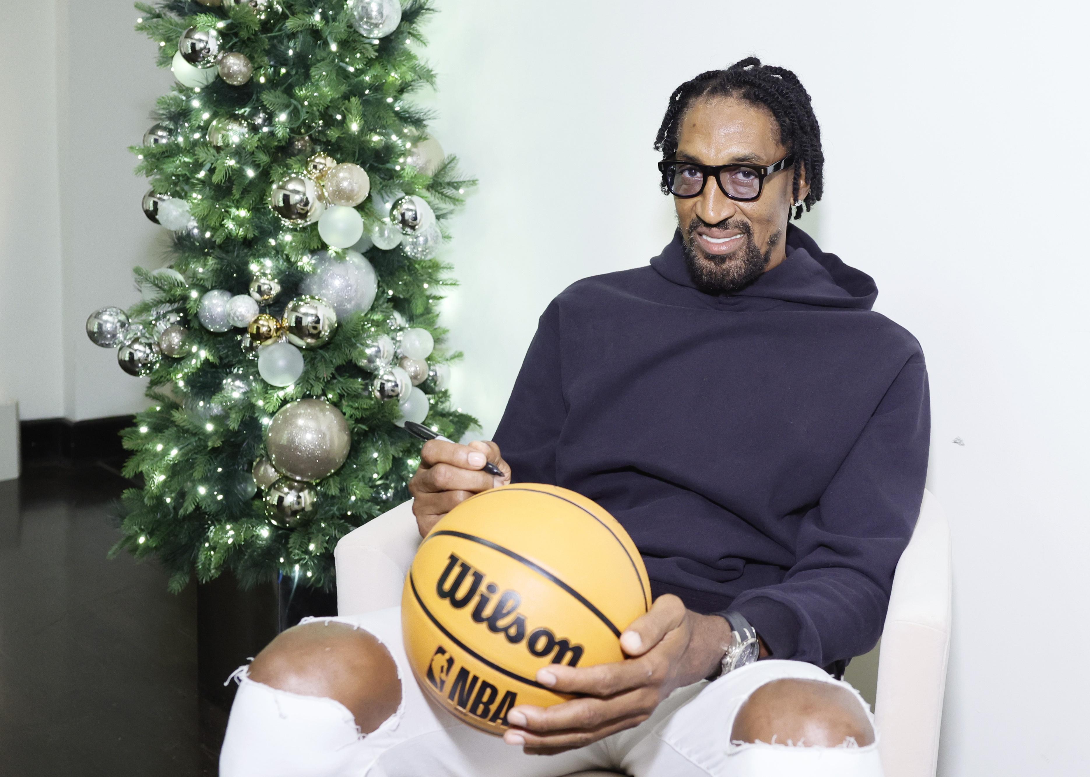 Scottie Pippen in a black hoodie and white jeans signing a basketball.