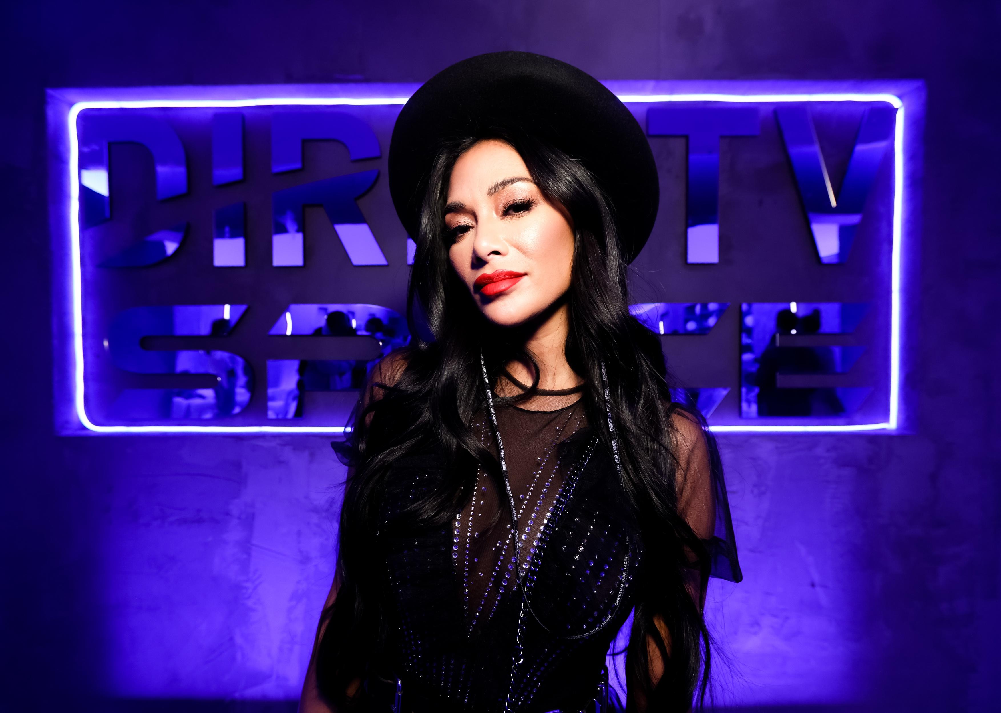 Nicole Scherzinger dressed in black with a black hat in front of a neon blue sign.