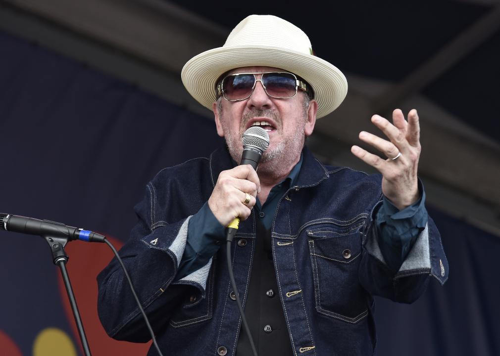 Elvis Costello performs in a dark denim jacket, hat and sunglasses.
