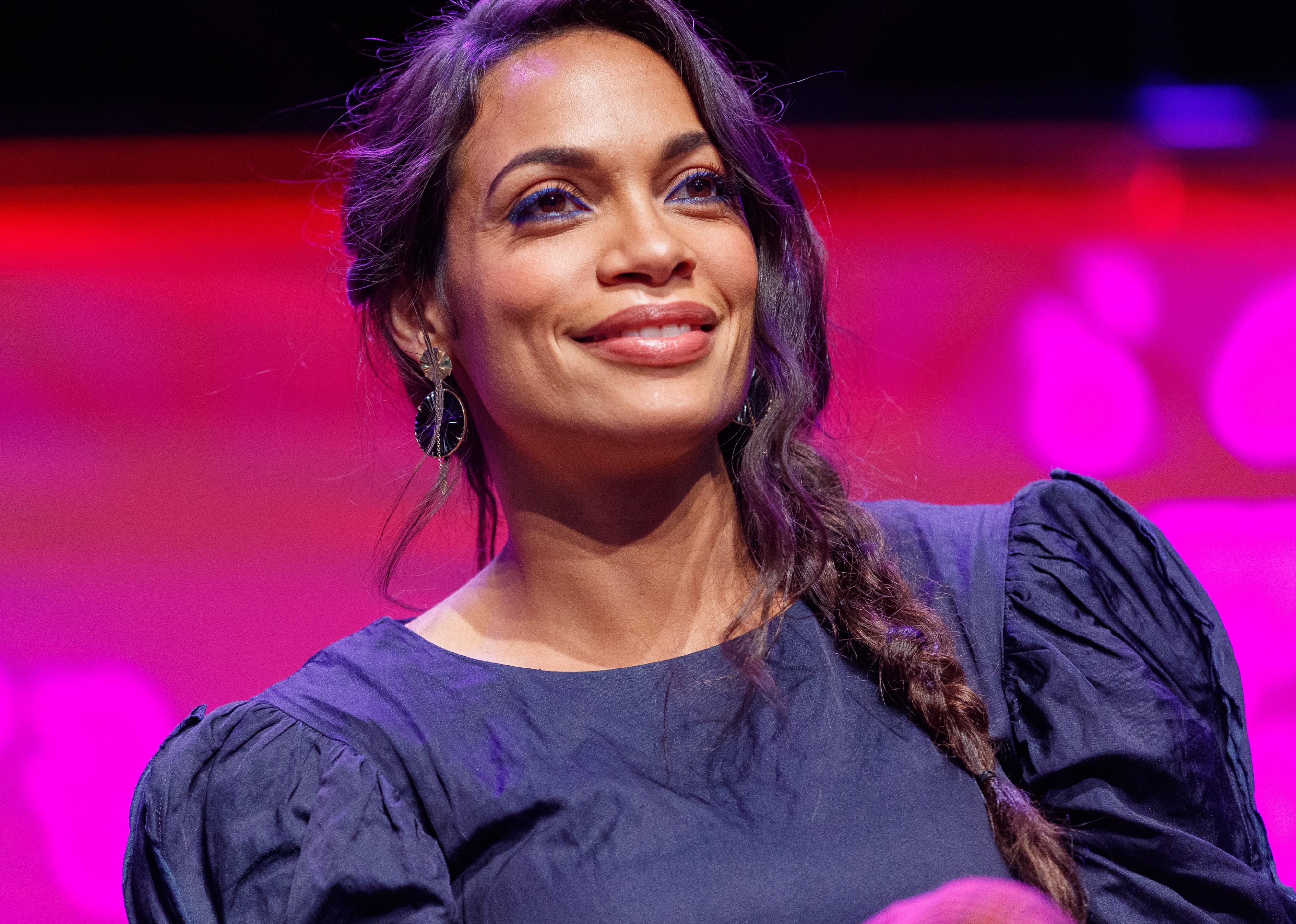 Rosario Dawson in black in front of a pink background.