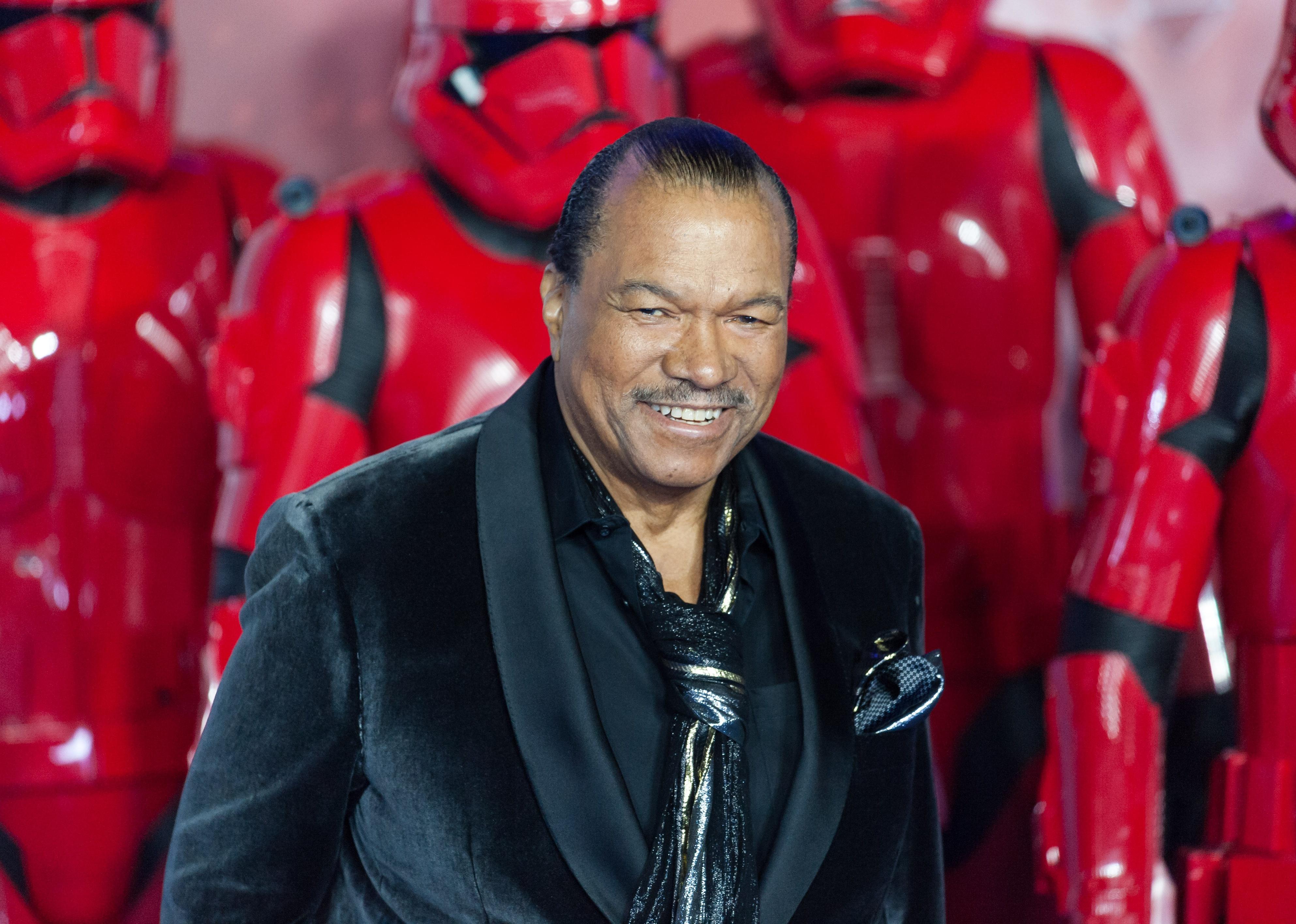 Billy Dee Williams on stage in a black velvet suit and scarf in front of red Star Wars suits.