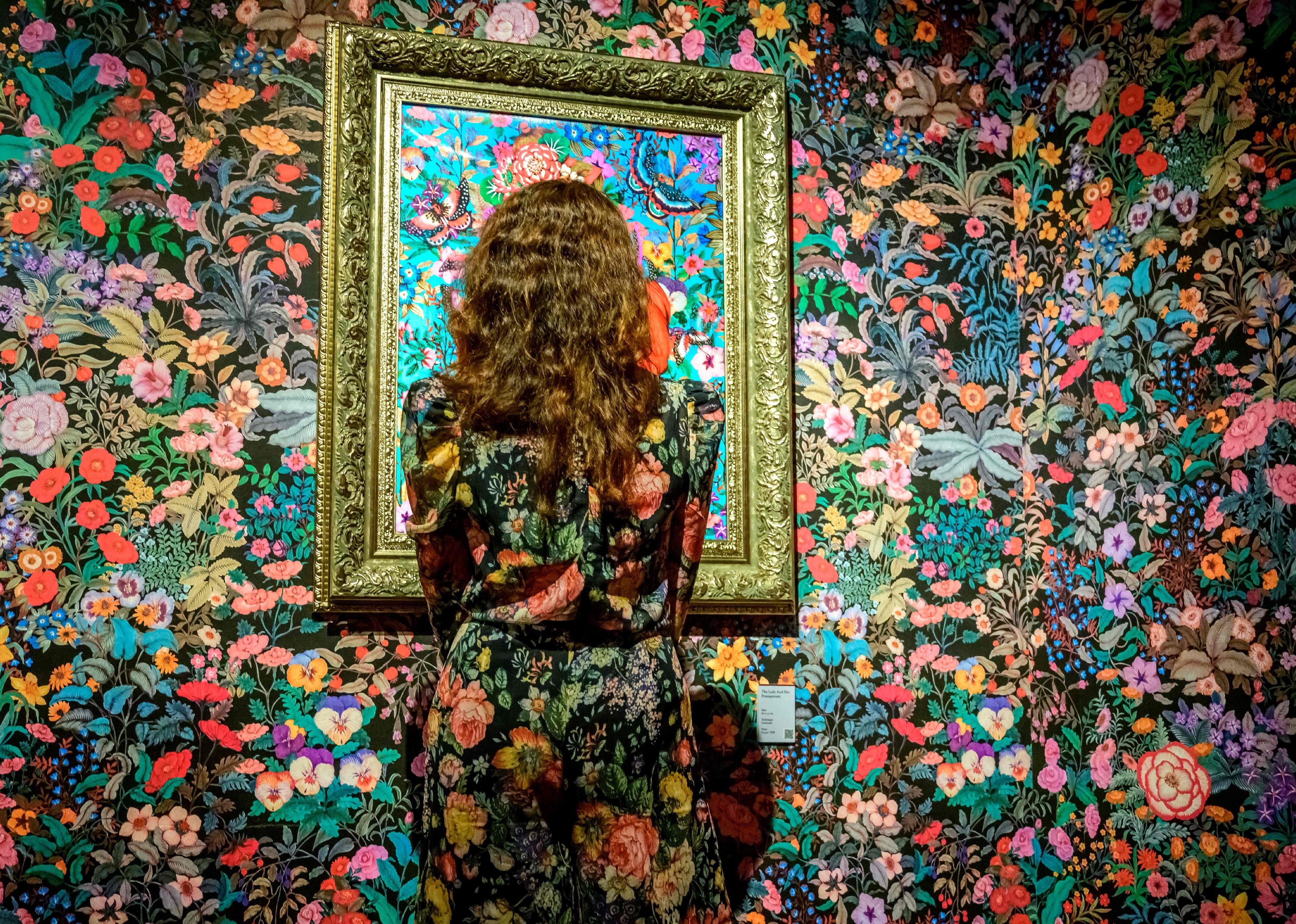 A woman dressed on floral print outfit looking at a painting by artist Phannapast "Yoon" Taychamaythakool that is surrounded by floral wallpaper.