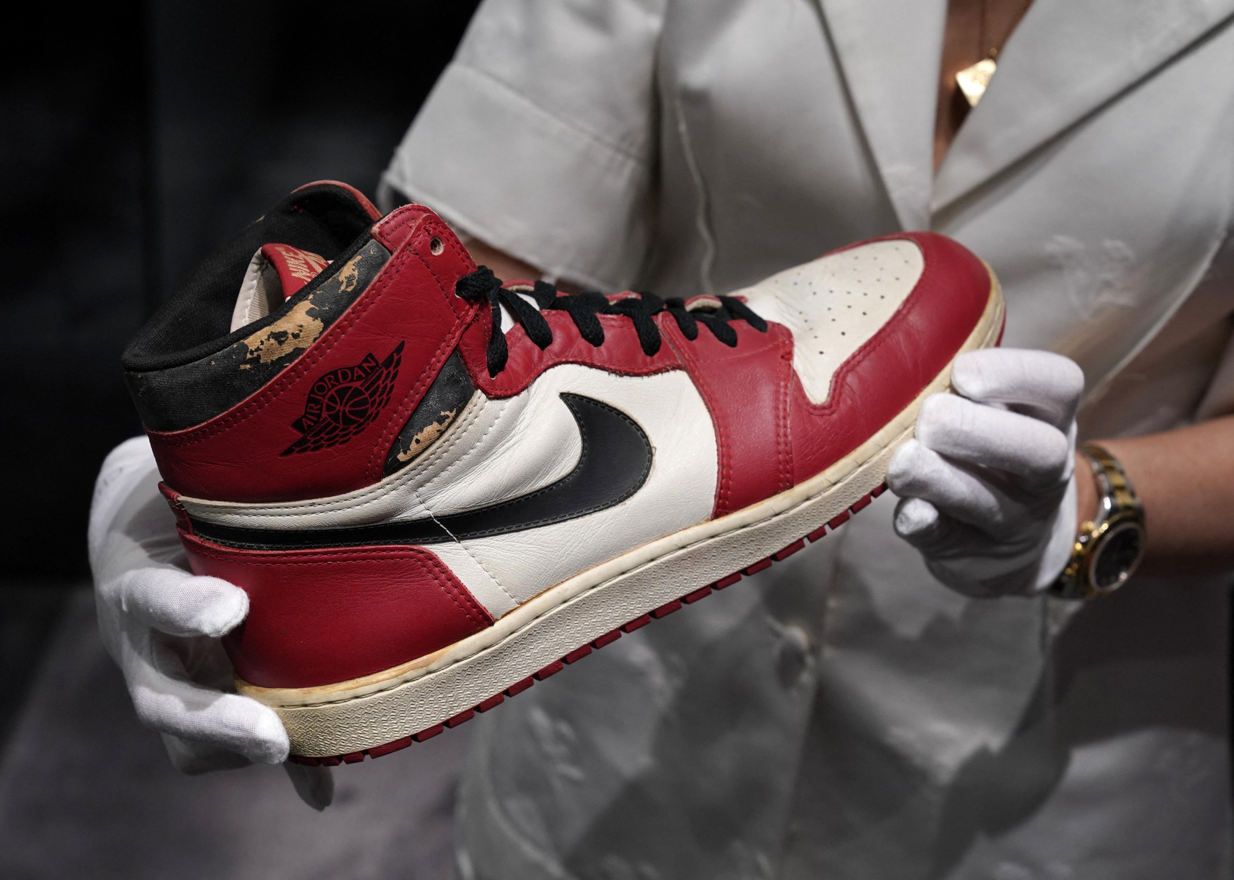A person wearing white gloves holds the Air Jordan 1 High Shattered Backboard Origin Story, Game-Worn Signed Sneaker Nike, 1985 Size 13.5, red, white and black.