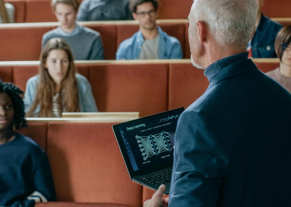 Professor holds a laptop while speaking in front of a college class.